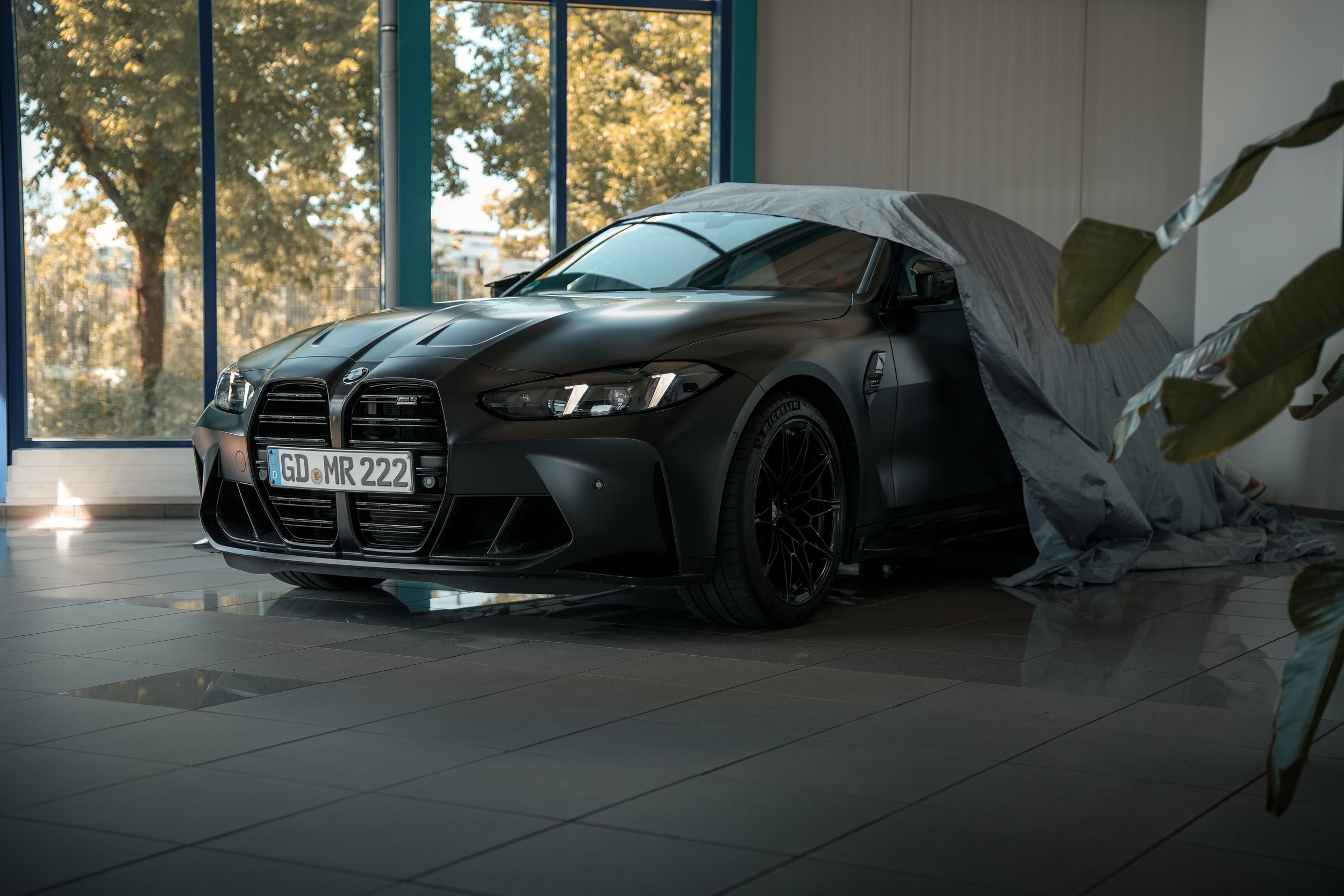 A black BMW M4 sports car inside a modern showroom with large windows and indoor plants, partially covered by a gray cloth.
