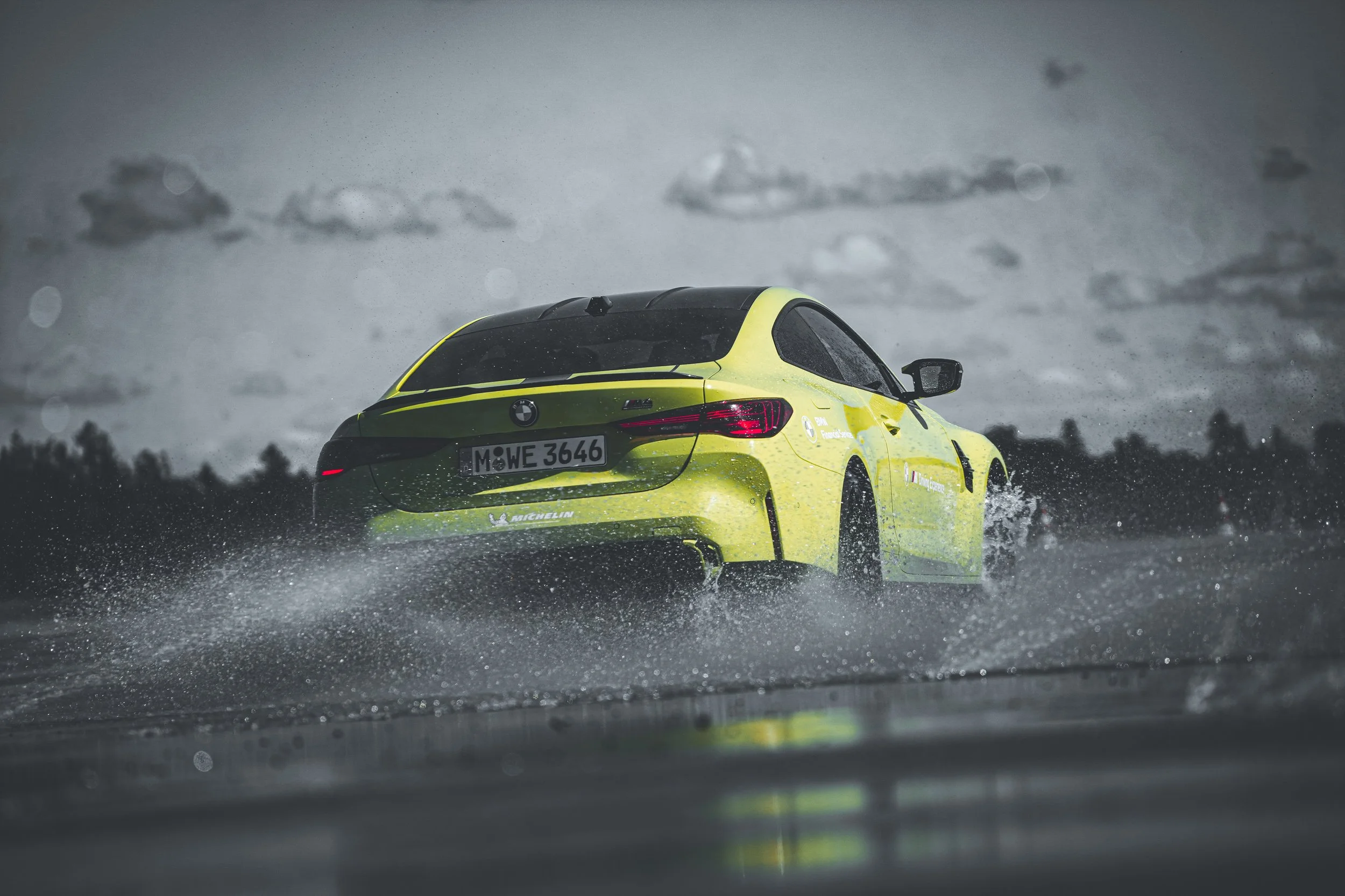 A yellow BMW M4 sports car driving through a rainy landscape with water splashing around and dark, cloudy sky in the background.