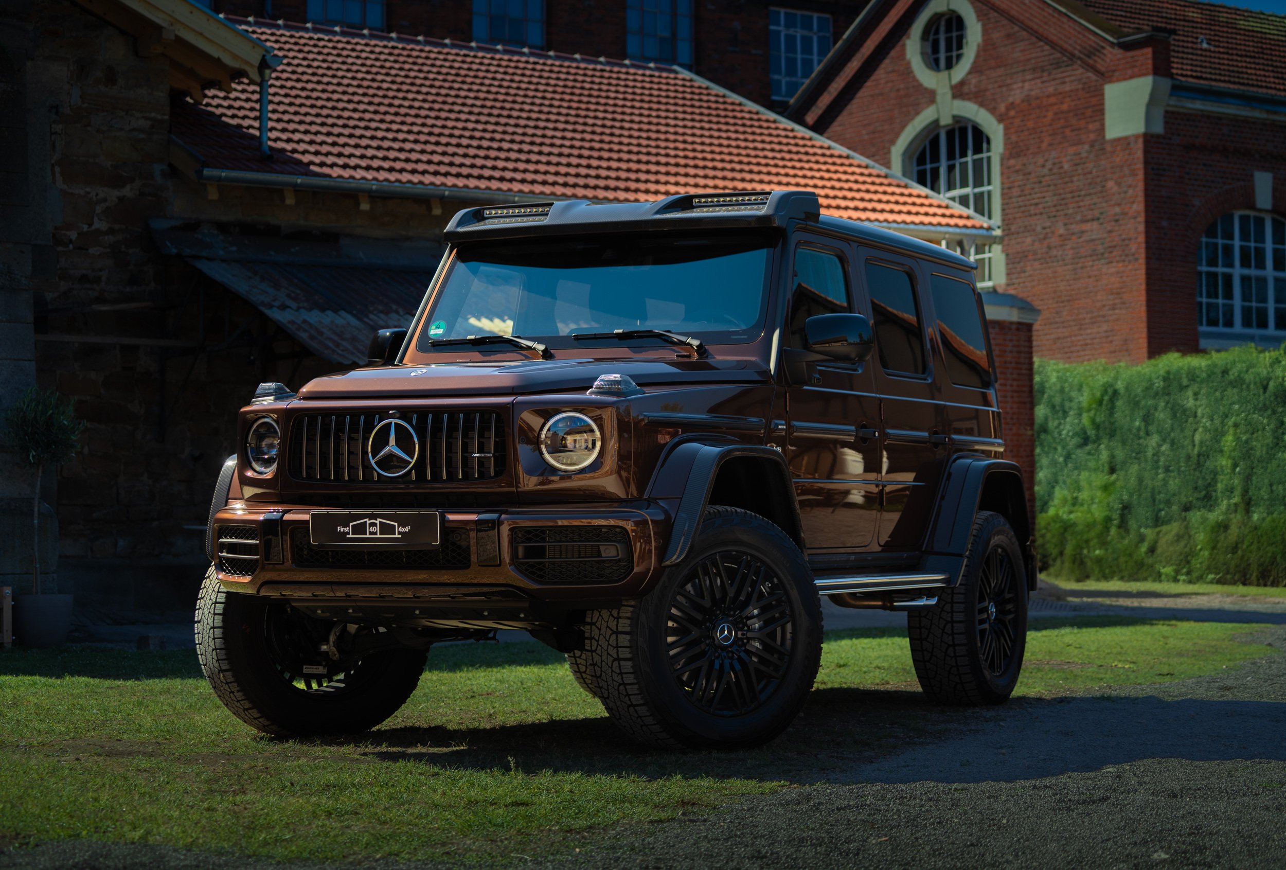 A black Mercedes-Benz G-Wagon parked on a green lawn with a brick house and a tiled roof in the background.