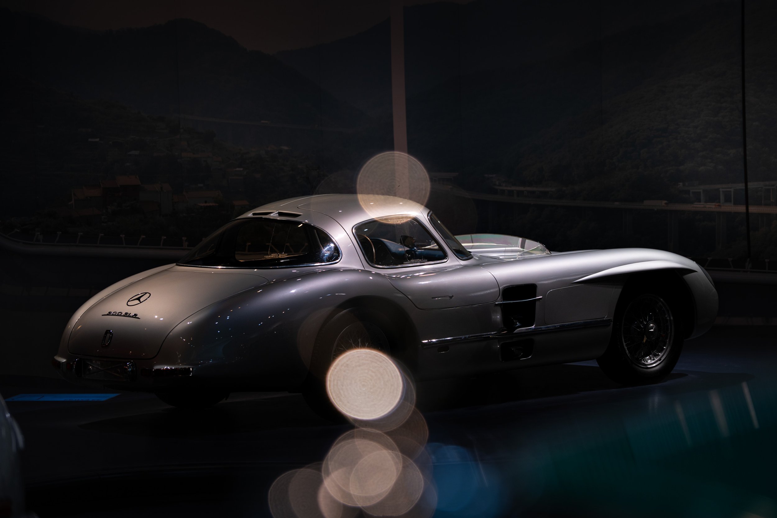 A silver Mercedes-Benz racing car displayed in a dimly lit indoor setting.