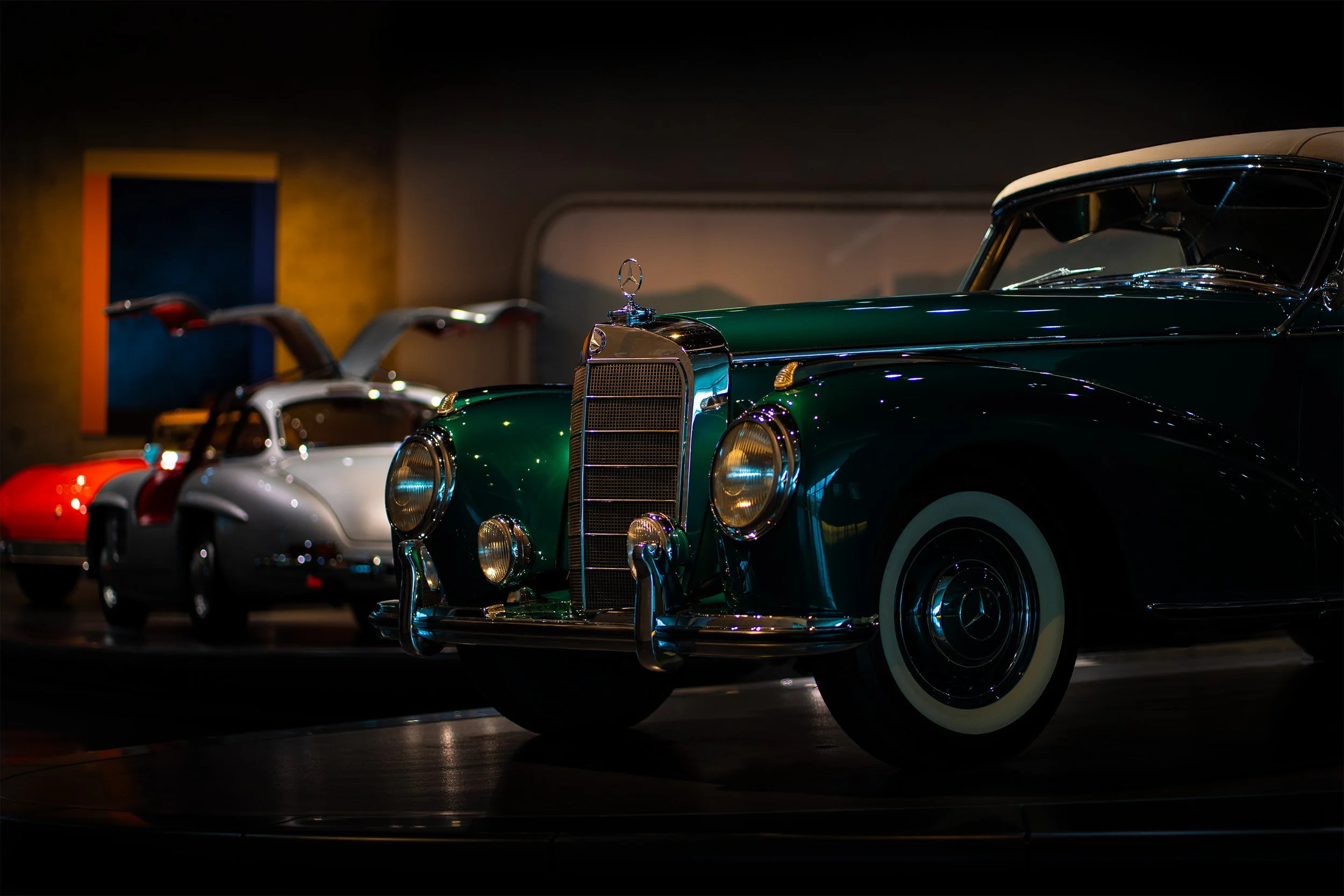 Vintage cars on display in a dimly lit showroom. The closest car is a green Mercedes-Benz with prominent chrome details, and there are two additional cars in the background, one red and one white, with their front ends visible.