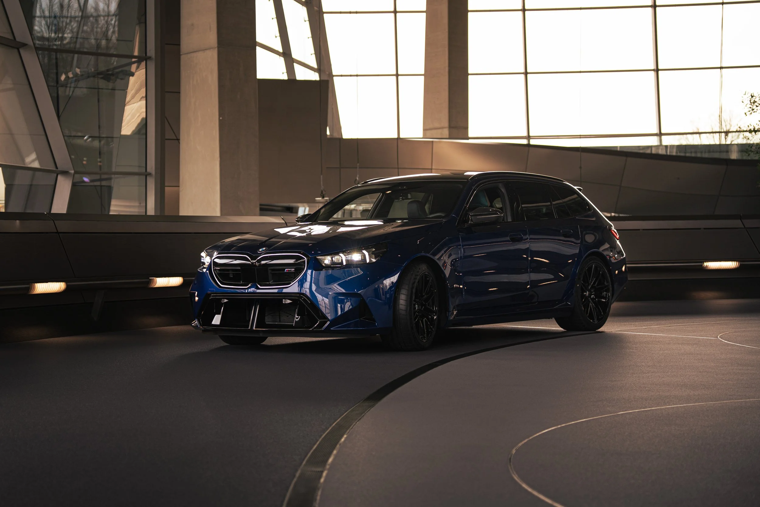 A blue BMW racing car with black wheels and a sleek design parked indoors at a modern building with large glass windows and concrete pillars, illuminated by natural light.
