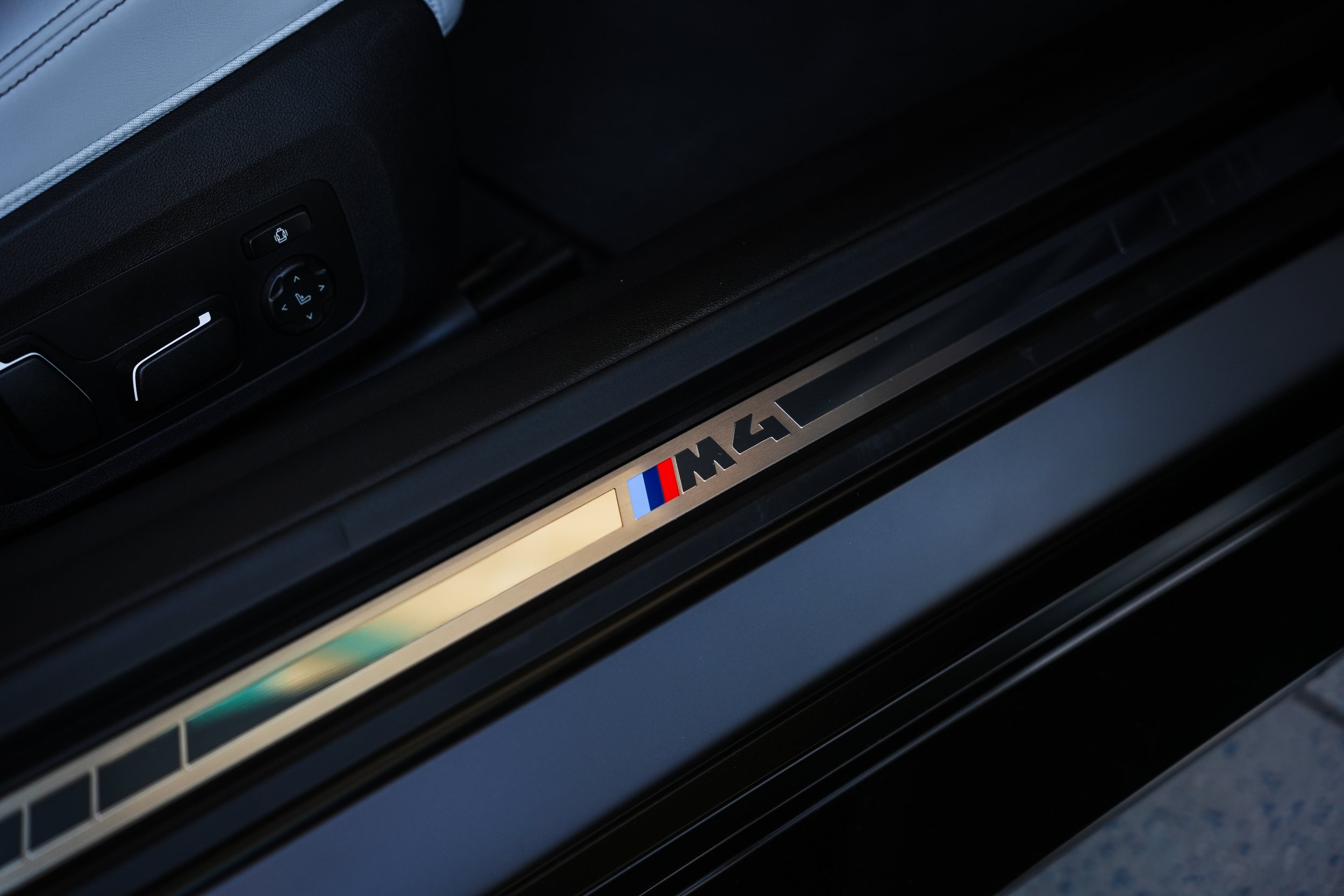 Close-up of a car interior door sill with a metal plate featuring the BMW M4 logo and a blue, purple, and red stripe, with part of the seat and control buttons visible.