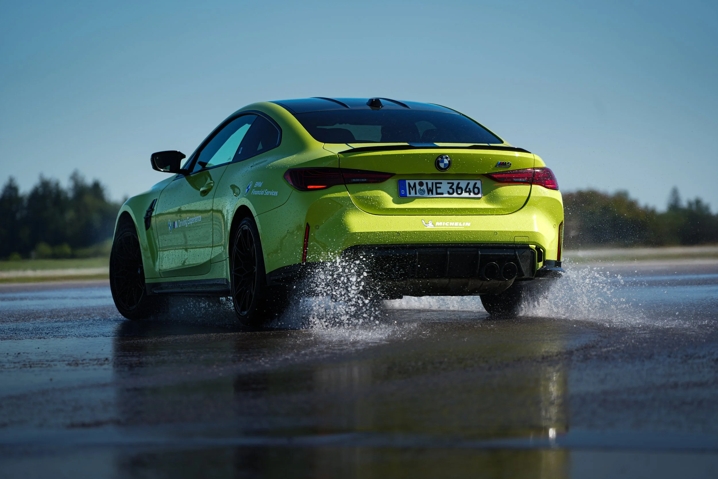 A lime green BMW M4 car driving through water, creating splashes, on a sunny day with clear skies and trees in the background.