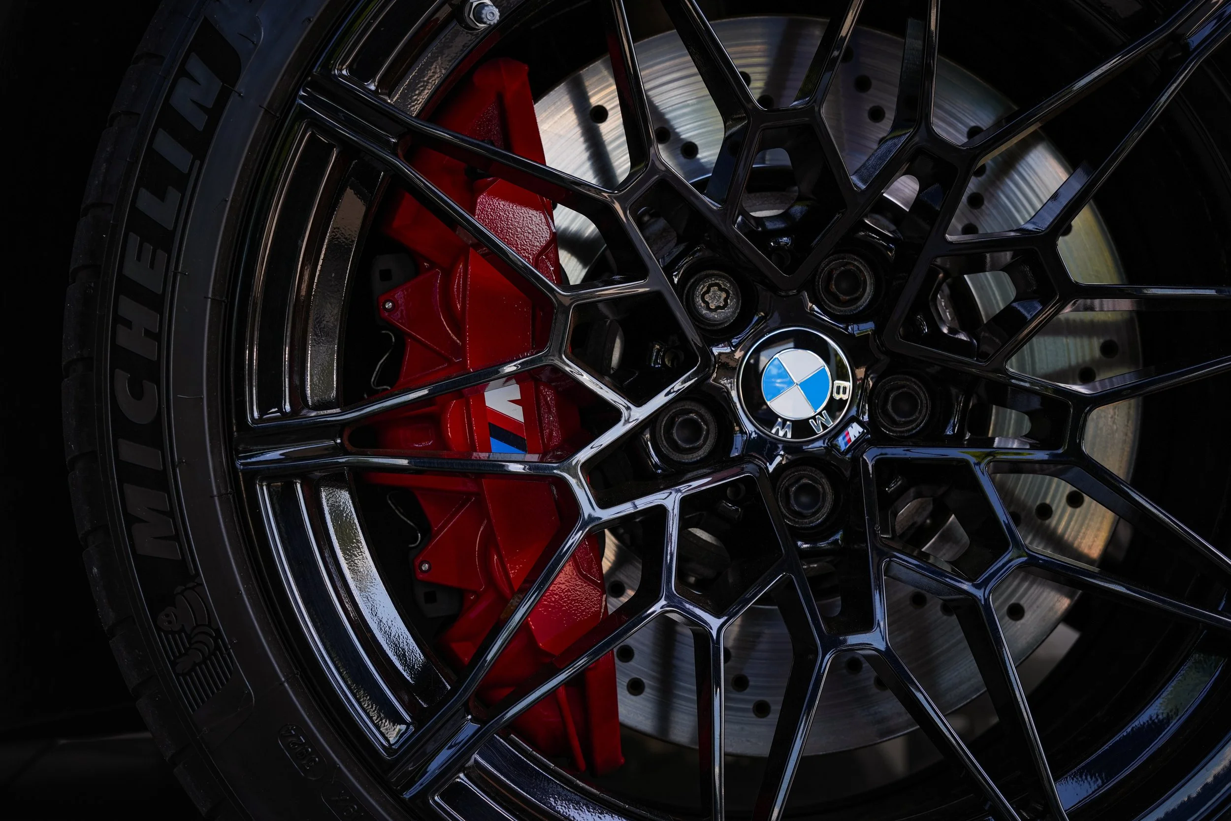 Close-up of a black alloy car wheel with a BMW logo at the center, showing red brake caliper and drilled brake disc behind the spokes.