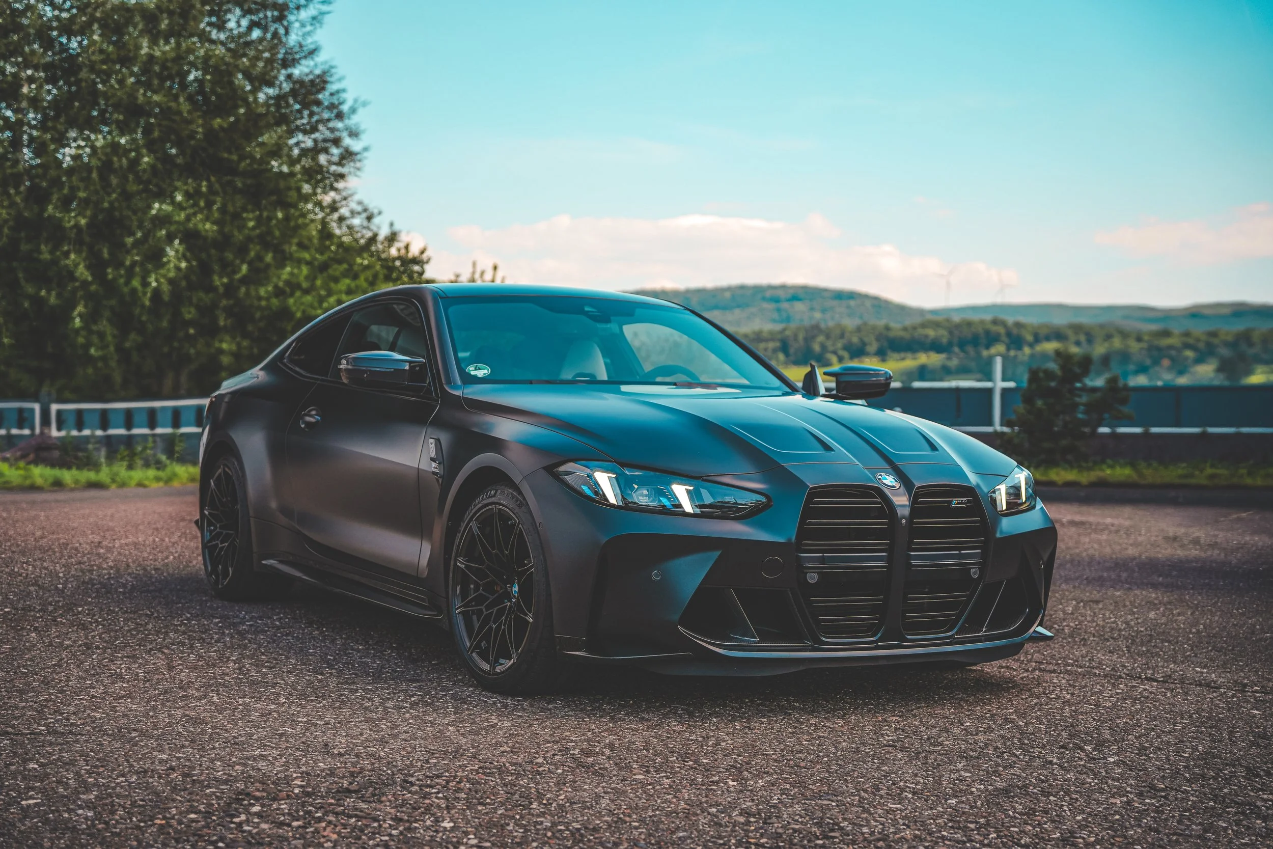 A matte black sports car parked on a gravel surface outdoors with a backdrop of trees, rolling hills, and a partly cloudy sky.