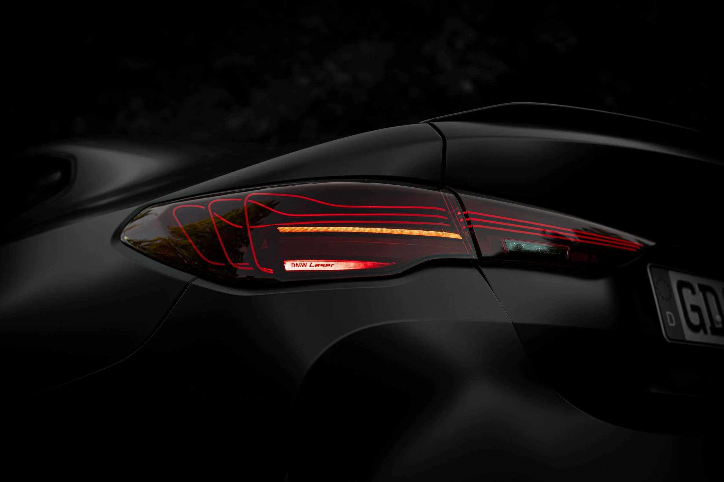 Close-up of the rear part of a black car showing a taillight with red and orange accents, with a reflection of trees on the car's surface.