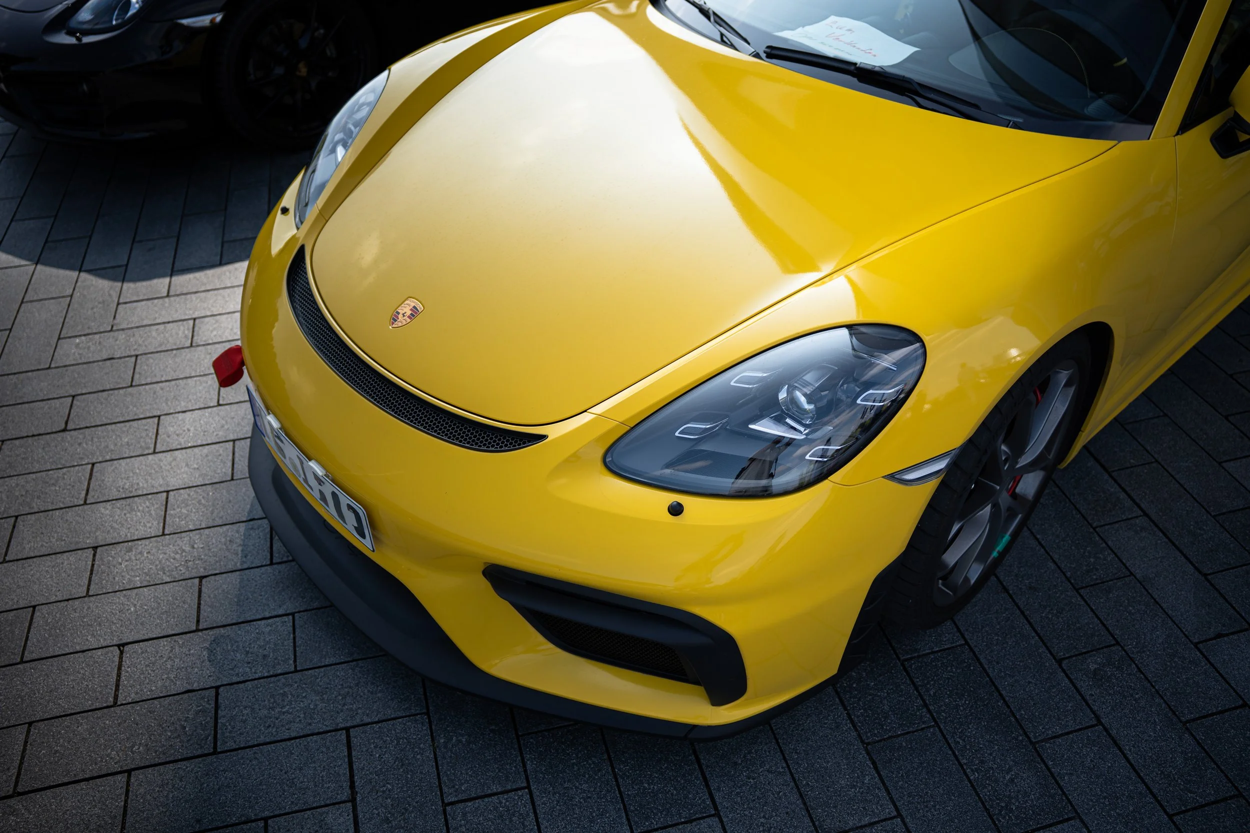 A yellow sports car with a Porsche logo on the hood, parked on a paved surface with another dark-colored car partially visible beside it.