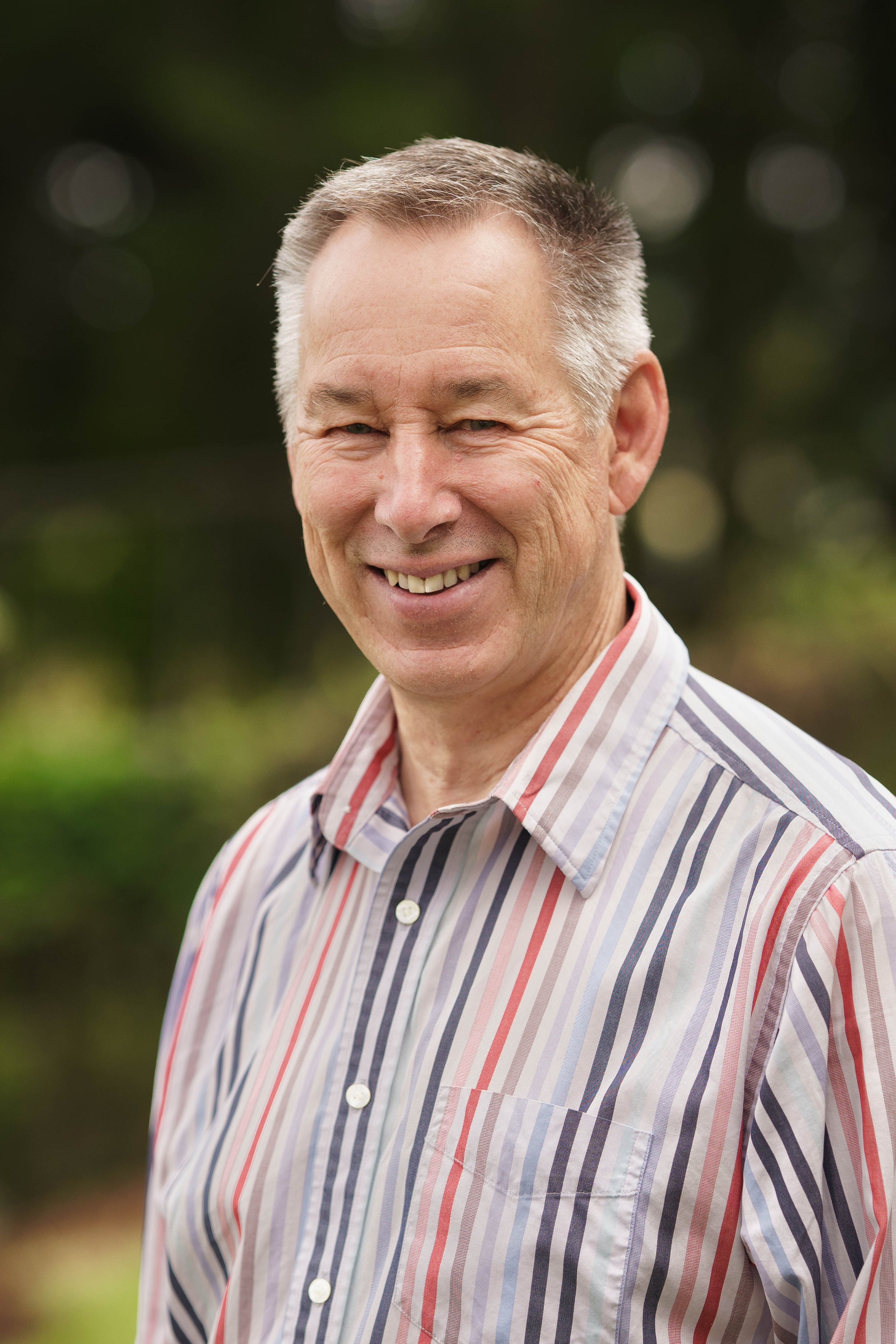 A smiling middle-aged man with short gray hair wearing a striped button-up shirt outdoors.
