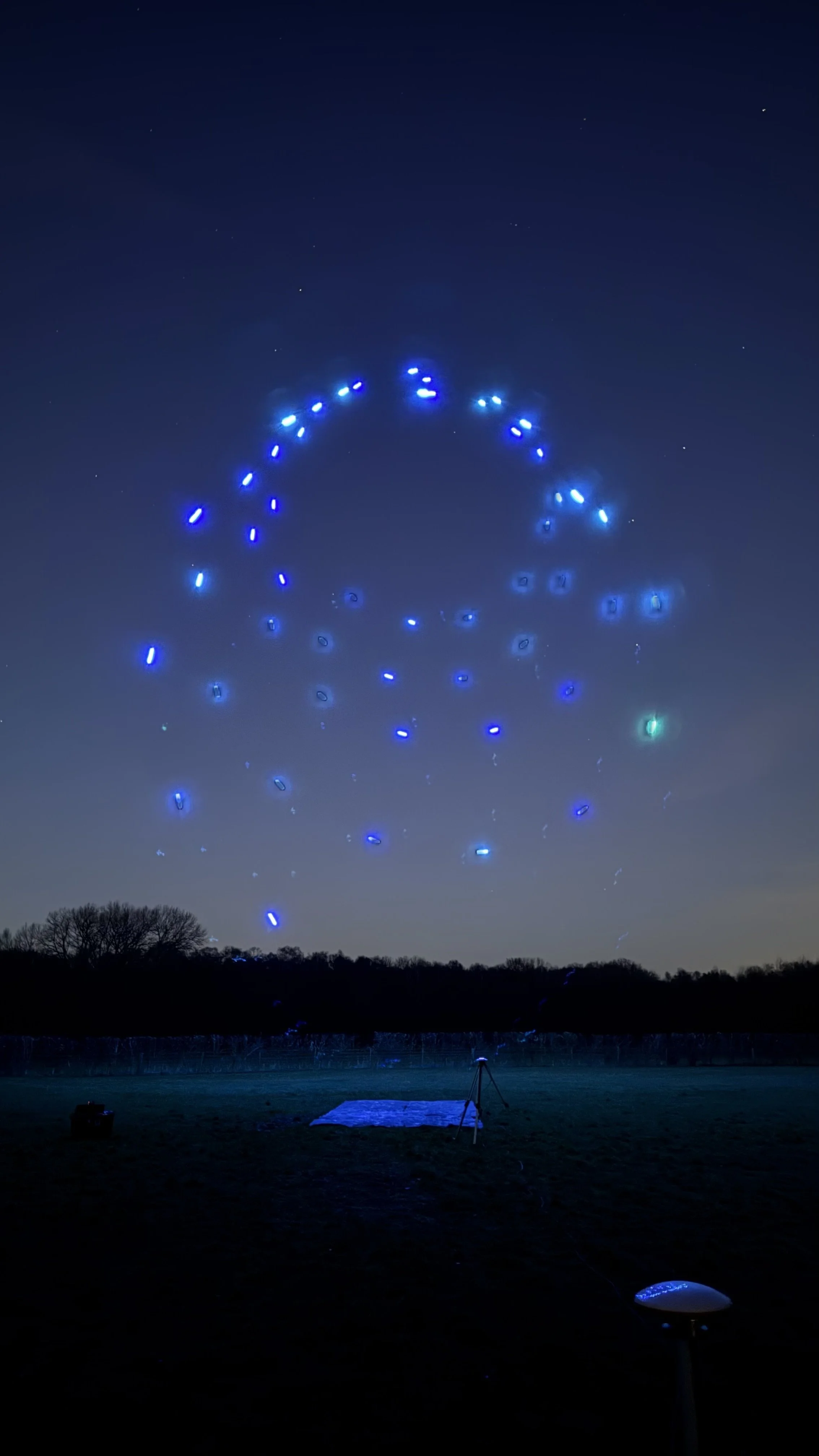 Night sky with a group of blue lights forming a circular pattern above a dark landscape, possibly drones in a light show.