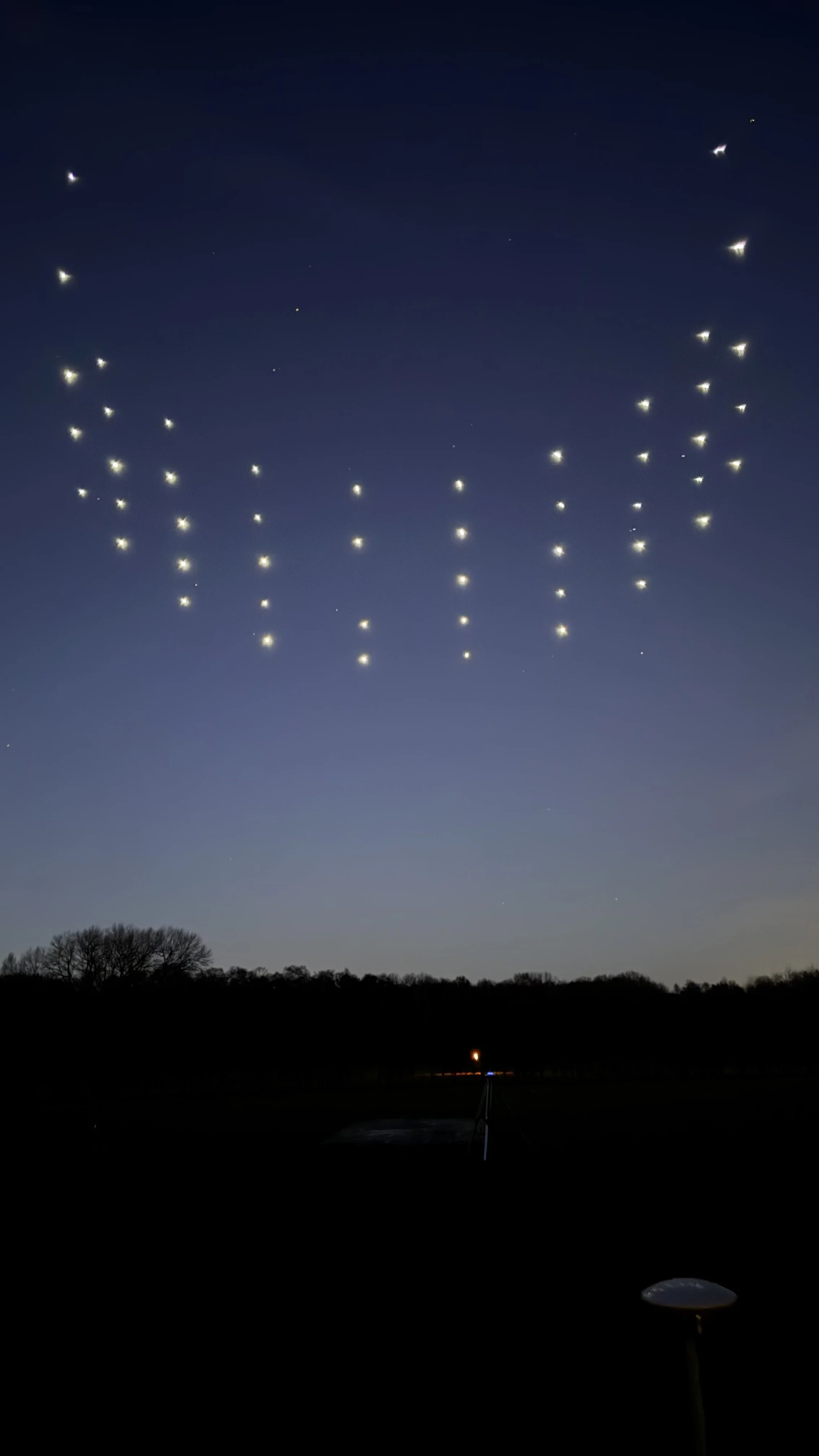 Drones with lights forming "WELCOME TO WORLD" in the night sky over a dark landscape with trees.