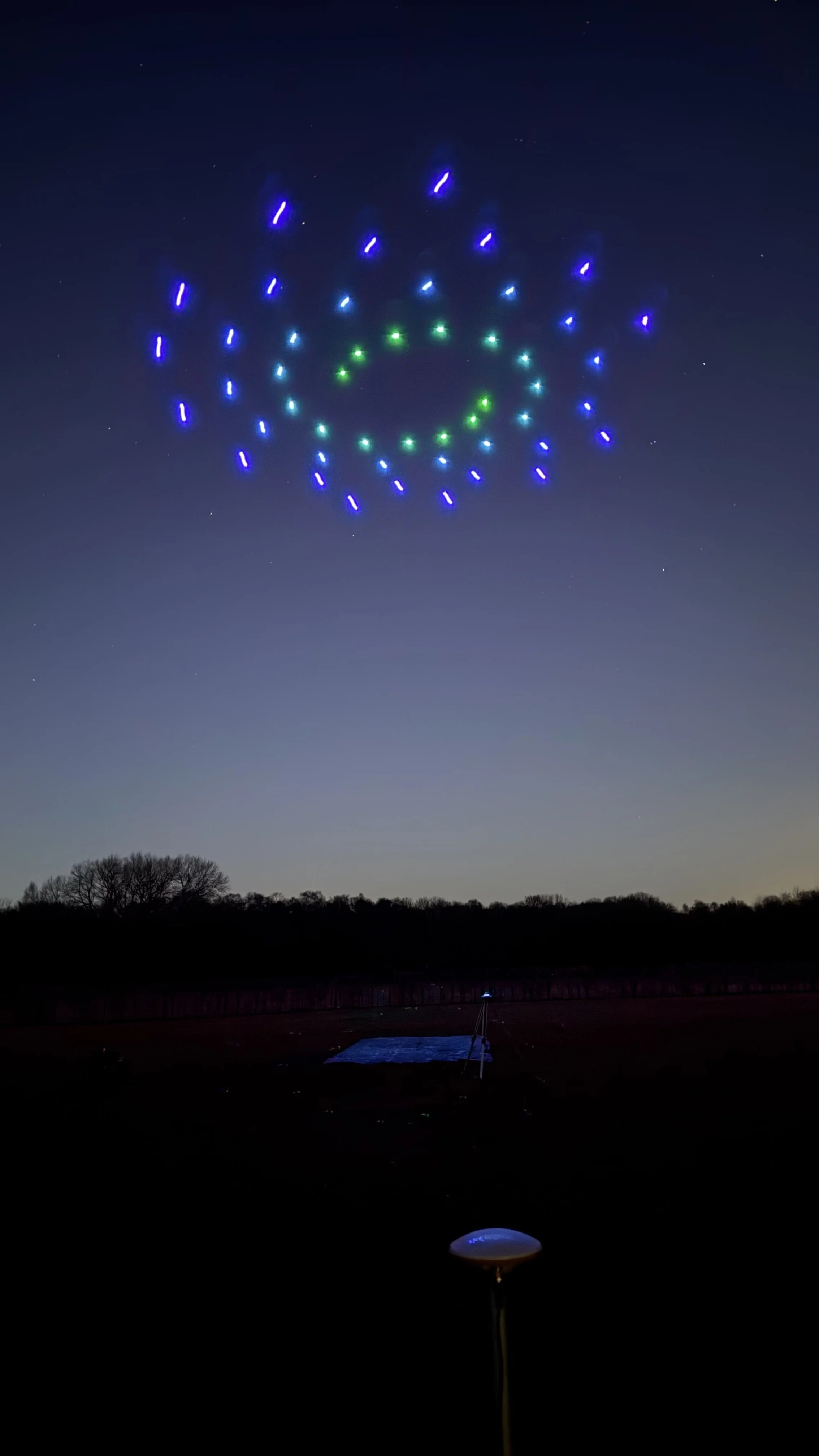 Drones with blue and green lights forming a circular pattern in the night sky, with trees in the distance.