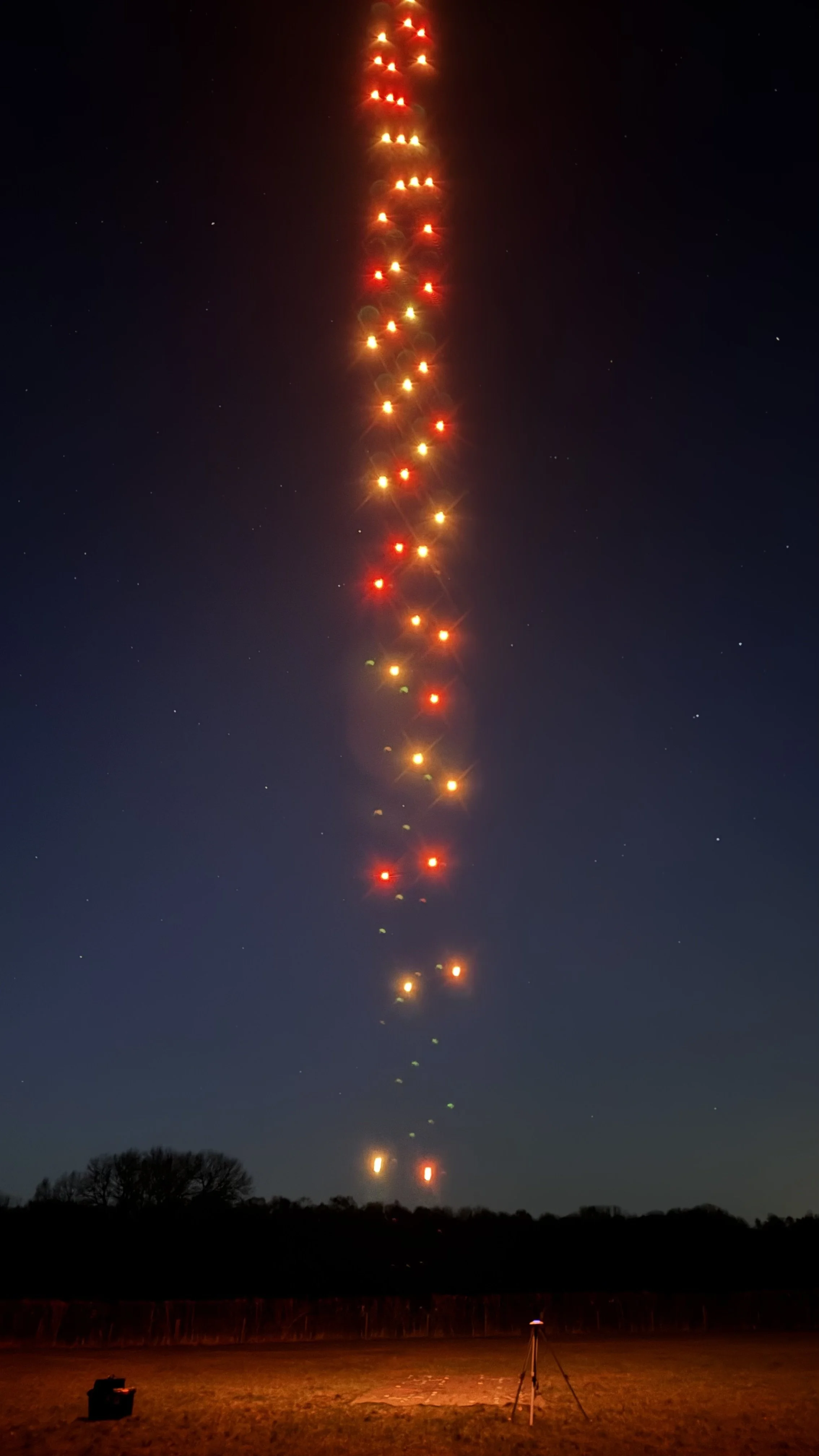 Long exposure photo of a rocket launch at night, showing a trail of bright, colorful lights in the sky with a tripod and equipment on the ground.