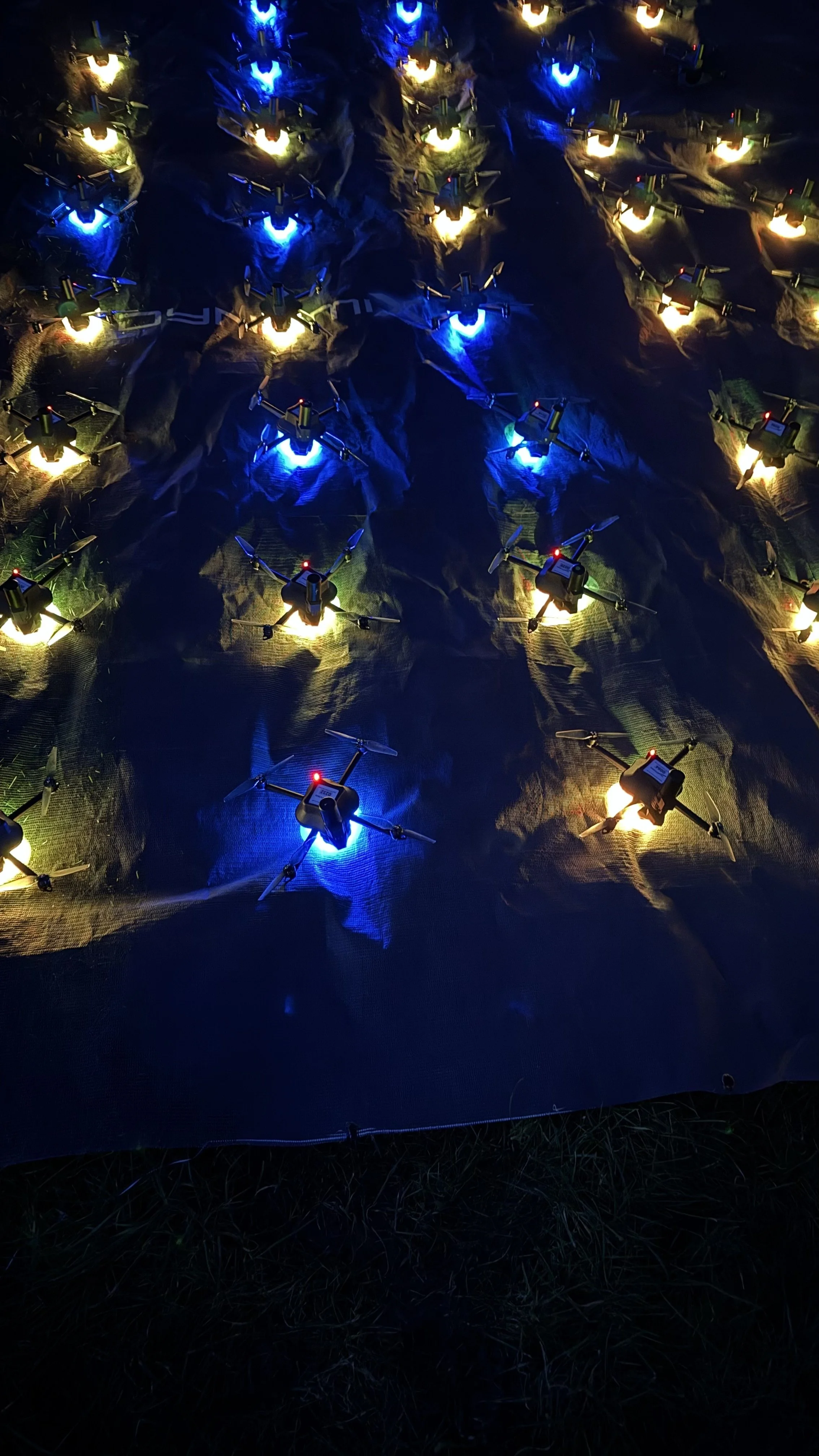Rows of illuminated drones with blue and yellow lights arranged on a dark surface at night.