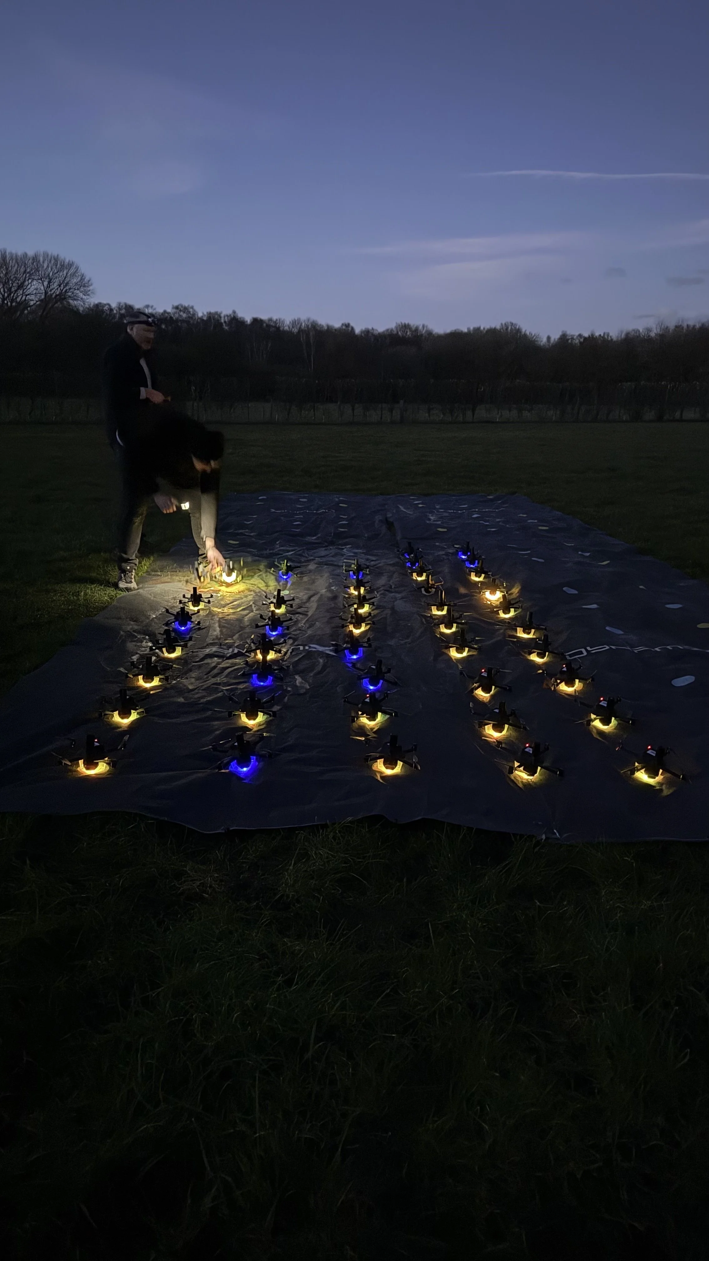 Person setting up illuminated drones on a dark field at dusk