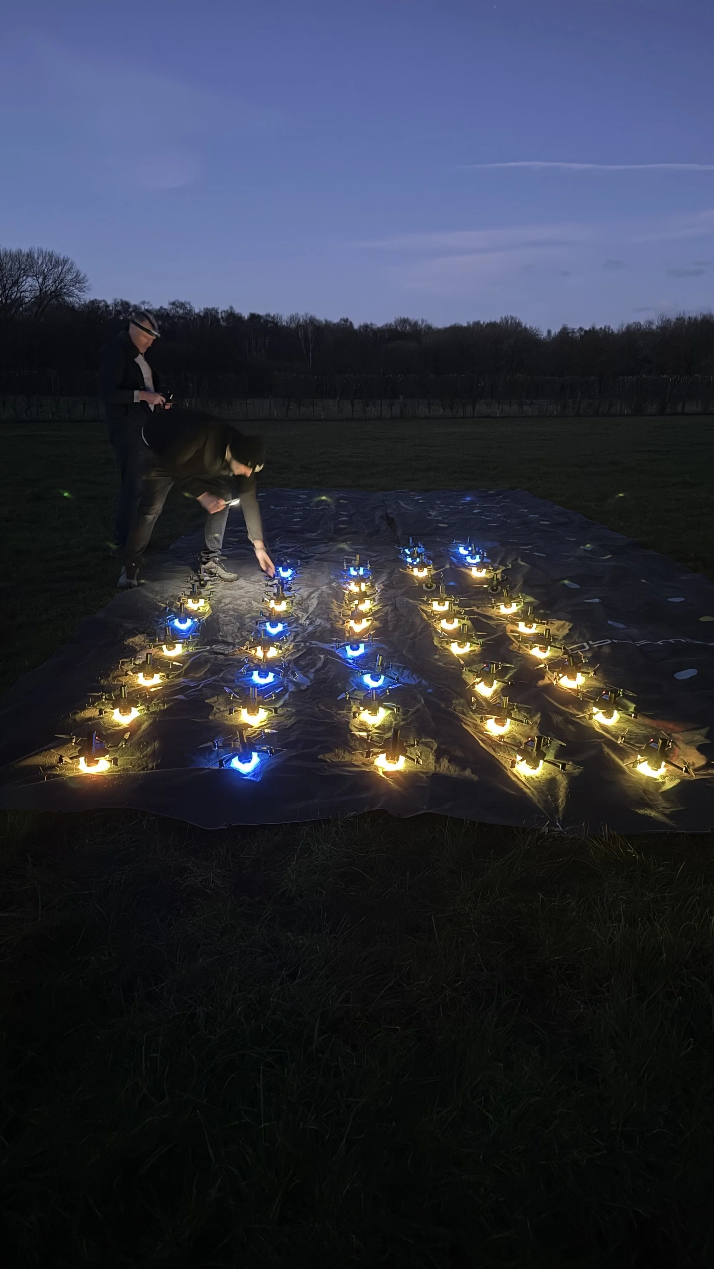 Two people arranging lights on the ground, glowing blue and yellow, in a field at dusk.