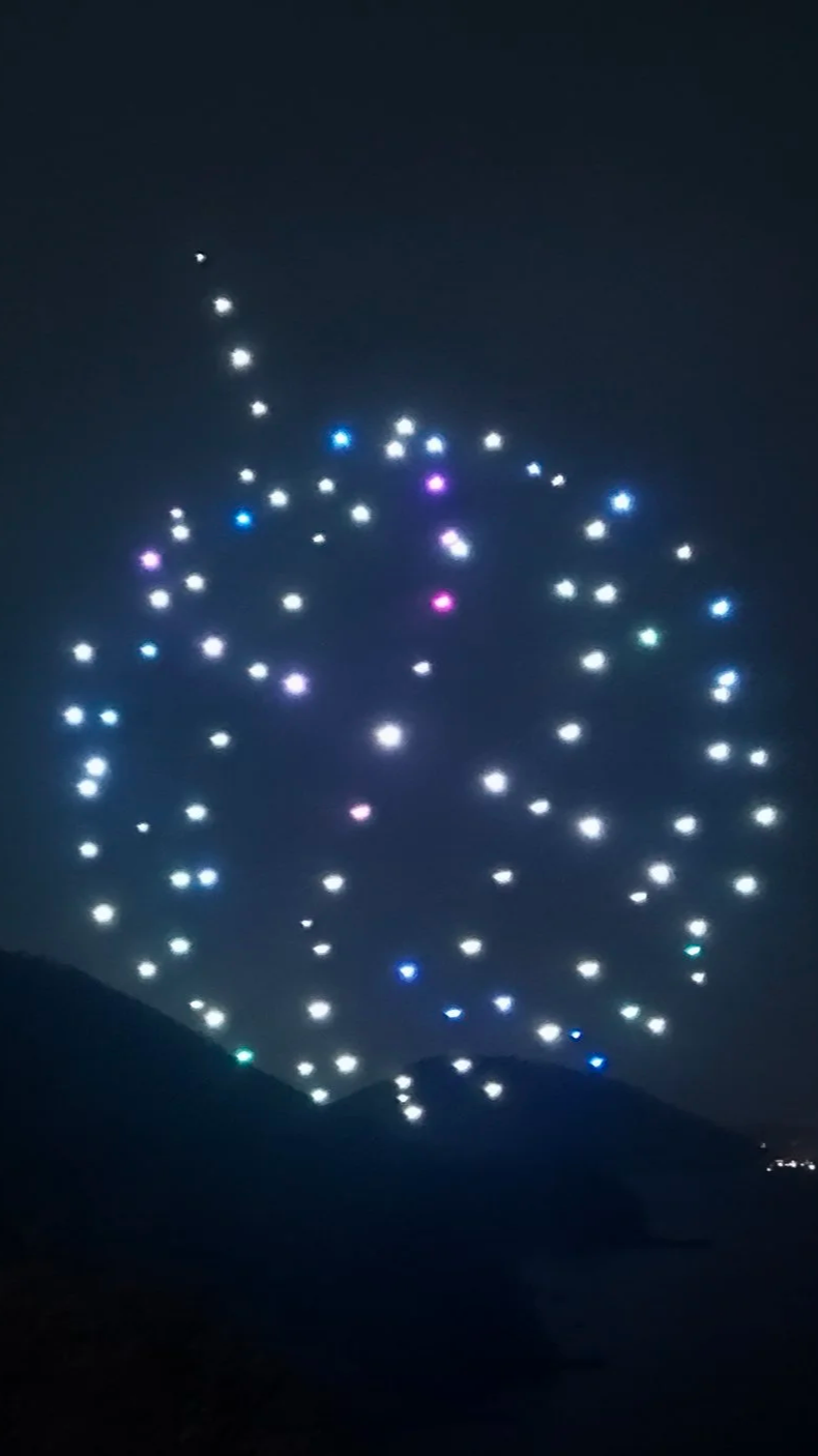 Night sky with a drone light show forming a large, abstract shape with colorful lights, above mountains.