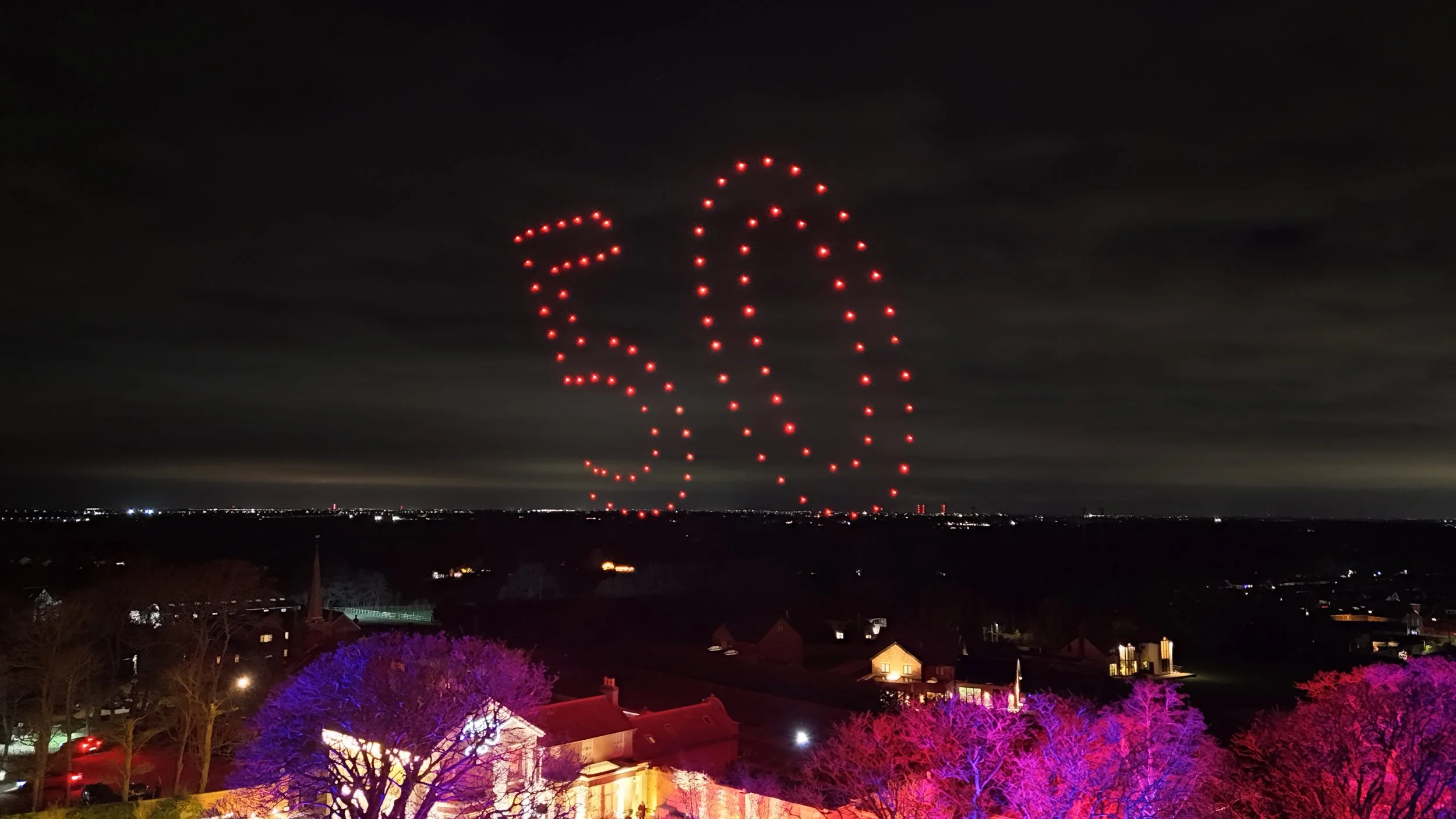 Nighttime scene with illuminated trees in pink and purple in the foreground and houses with lit windows. In the sky, a drone light display shows the number 30 in red lights against dark clouds.
