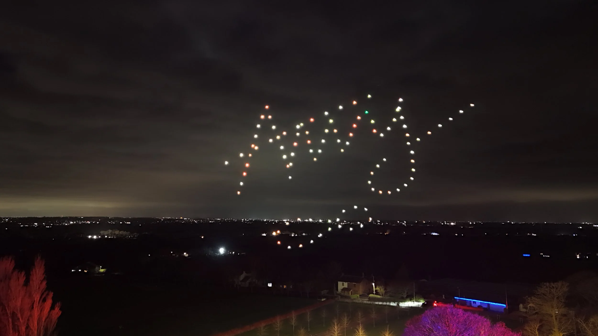 Night sky with drone lights spelling out '2023' and a star-shaped pattern, overlooking a dark landscape with few lights and trees illuminated in colorful lights.