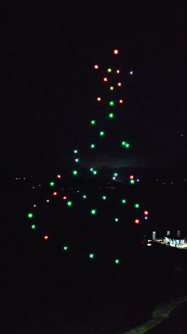 Night sky with a vertical string of colorful lights in red, green, blue, and yellow, arranged in a pattern resembling a Christmas tree.