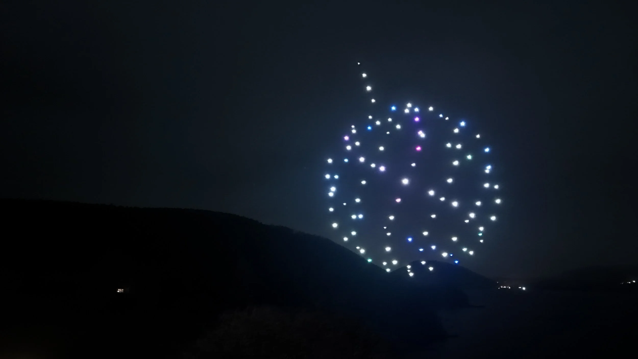 Nighttime scene with a large firework resembling a skull exploding in the sky, with colorful dots of light against a dark sky and silhouetted landscape.