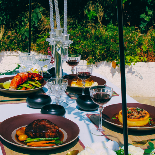 A table set for a meal outdoors with wine glasses, candles, and plates of food including steak, vegetables, and a lobster.