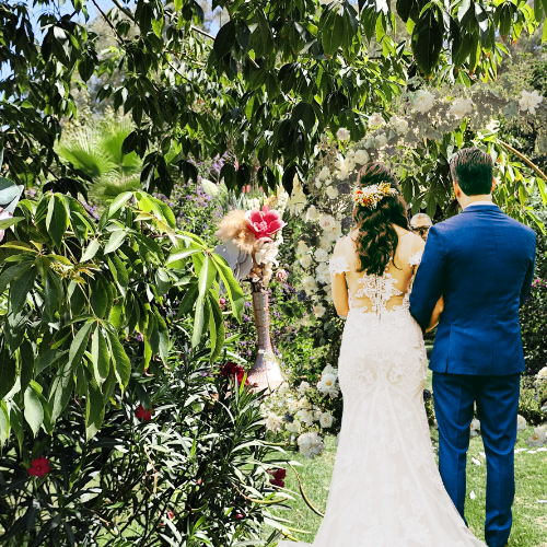 A bride and groom stand outdoors under a floral arch during their wedding ceremony, surrounded by lush greenery and flowering plants.