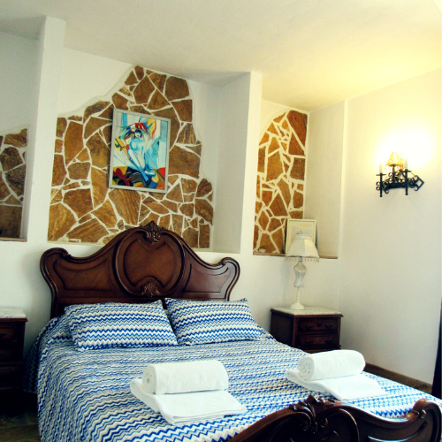 A bedroom with a wooden bed frame, blue and white bed linens, two pillows, and two rolled towels on the bed. There are two nightstands with lamps, a colorful artwork on a stone accent wall, and a wall-mounted light fixture.