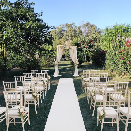 Outdoor wedding setup with white chairs arranged on both sides of a white aisle, leading to a decorated arch with white curtains and flowers, in a garden with trees and flowering bushes. Dragonfly retreat Spain