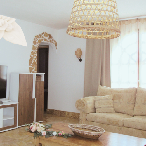 Living room with beige sofa, wicker pendant light, floral arrangement on table, stone archway, and curtain-covered window.