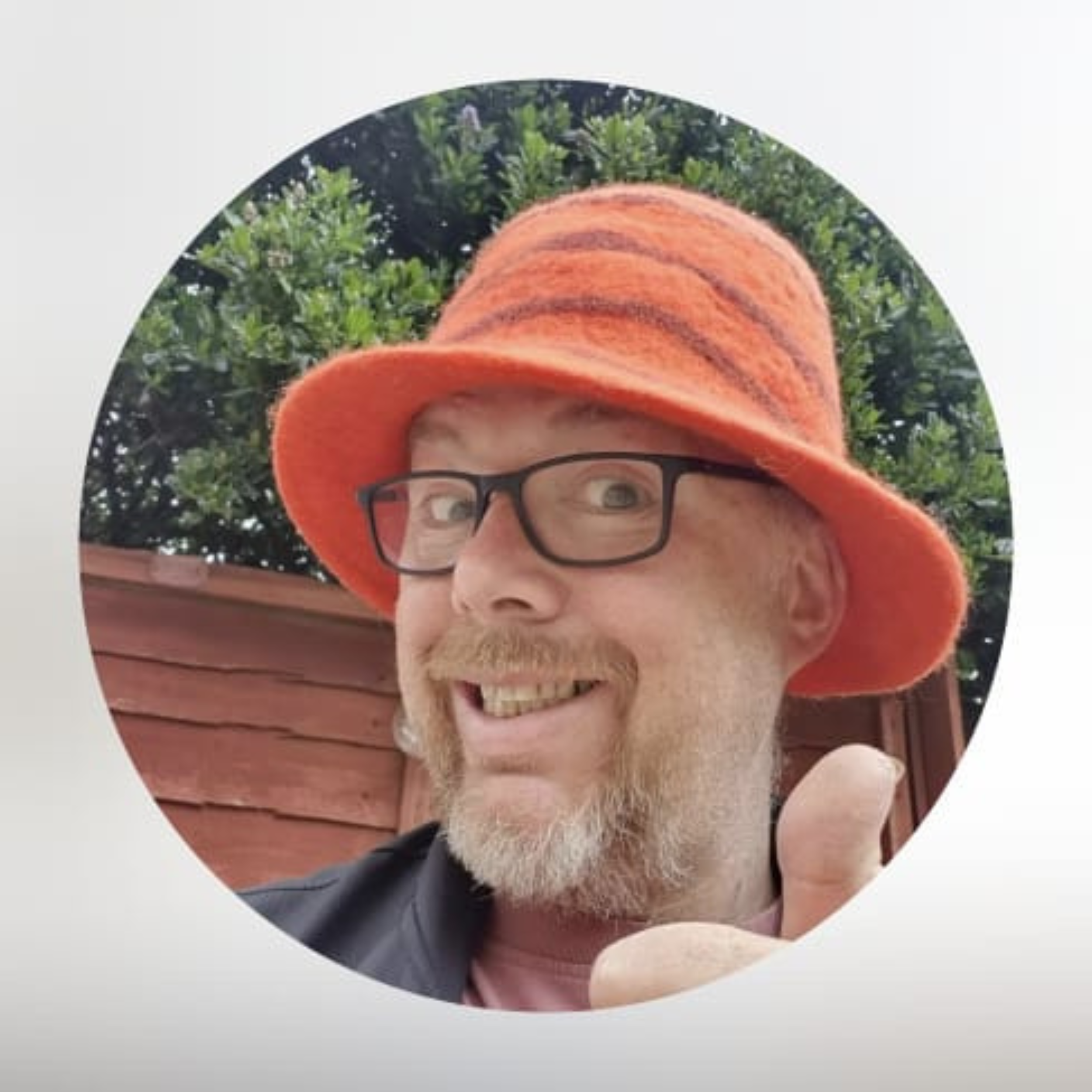 A man with glasses smiling and giving a thumbs-up, wearing an orange felt hat, outdoors with green trees and a wooden fence in the background.