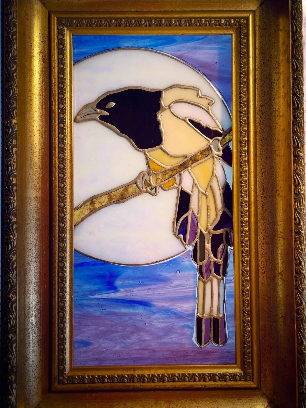 Stained glass window depicting a bird perched on a branch with a large moon or sun in the background, framed by ornate gold frame.