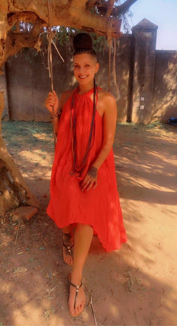 A woman wearing a sleeveless red dress, black sandals, and long necklaces, standing outdoors beside a tree with a swing. She has dark hair styled in a structured bun and is smiling.