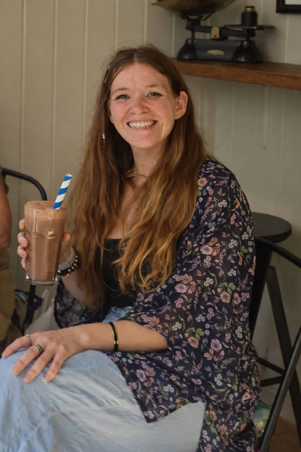 A young woman with long red hair, smiling, holds a glass of chocolate milkshake with a blue and white striped straw, sitting indoors with a beige wall and a shelf behind her.