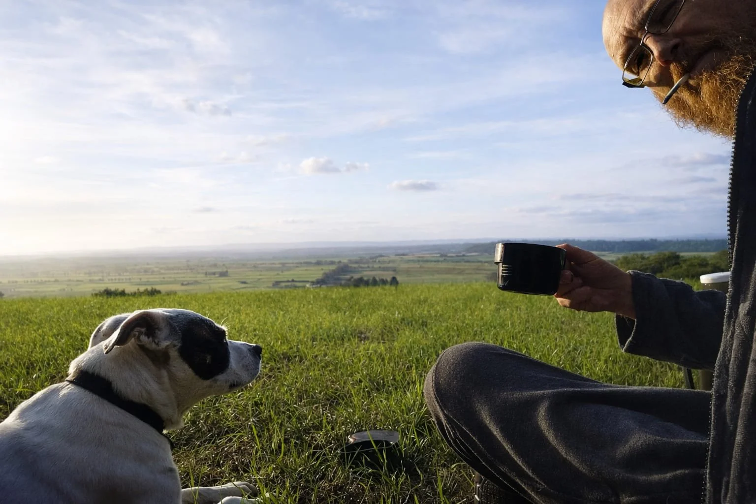 A man with a beard and glasses sitting in a grassy field with a dog, holding a black mug, with a view of a wide landscape and blue sky in the background.
