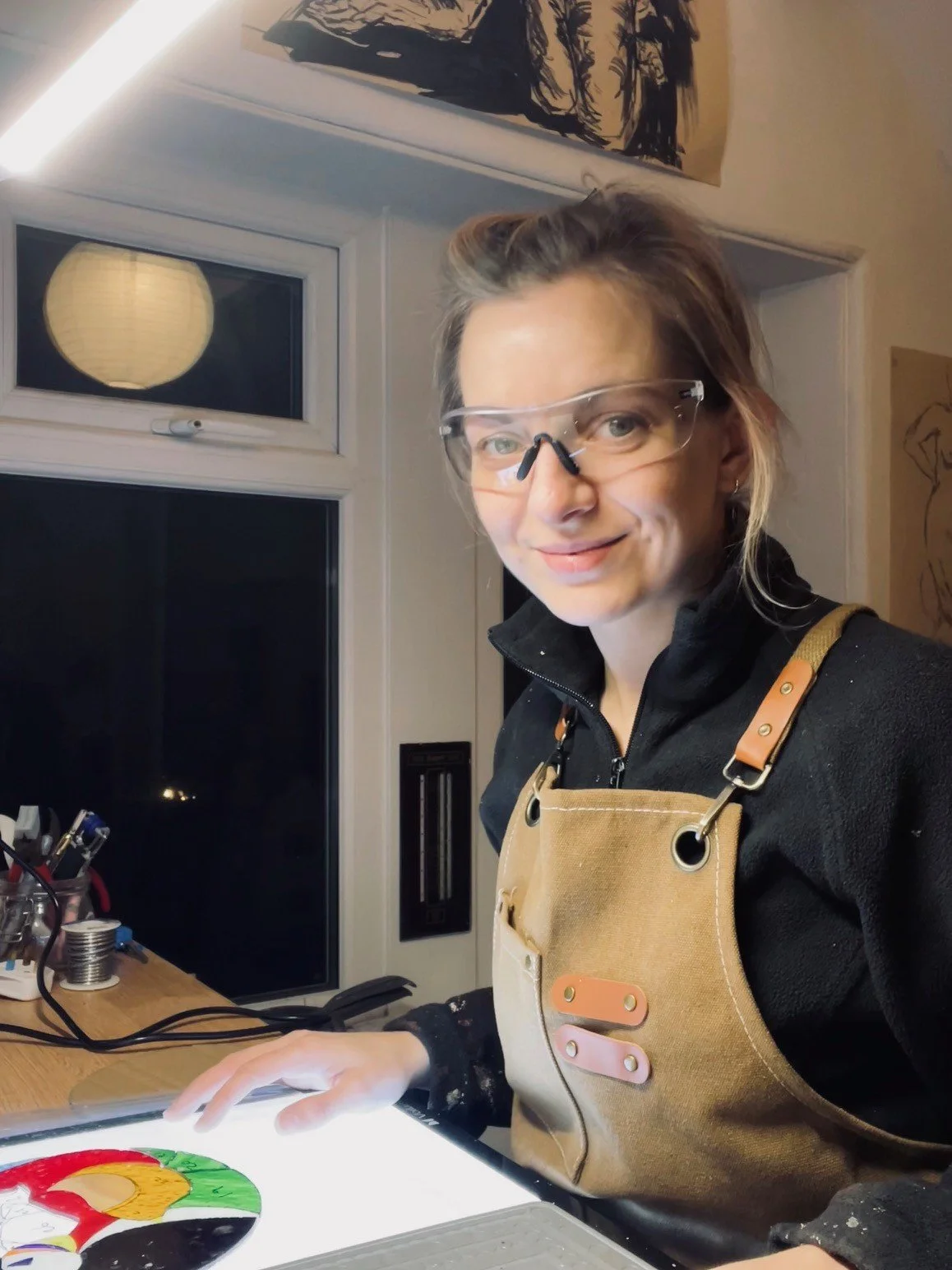 A woman with glasses and a light brown apron sitting at a desk in a room, working on a colorful art project with a circular palette, with window and paper lantern light in the background.