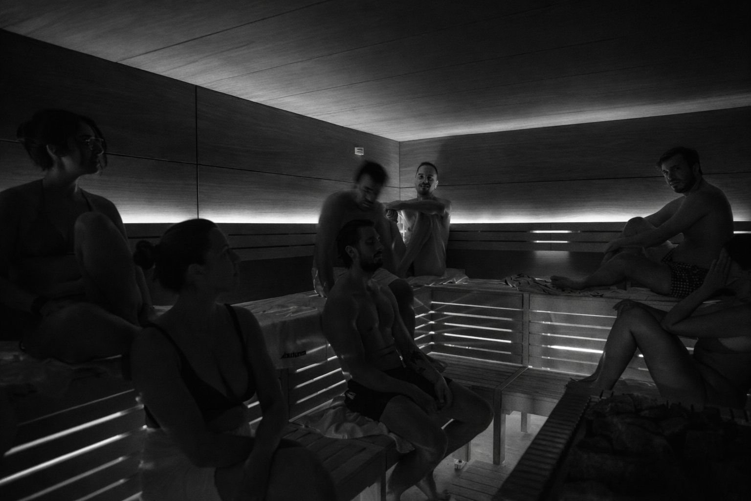 Grupo de pessoas em uma sauna com iluminação suave, algumas sentadas e outras de pé, conversando e relaxando.