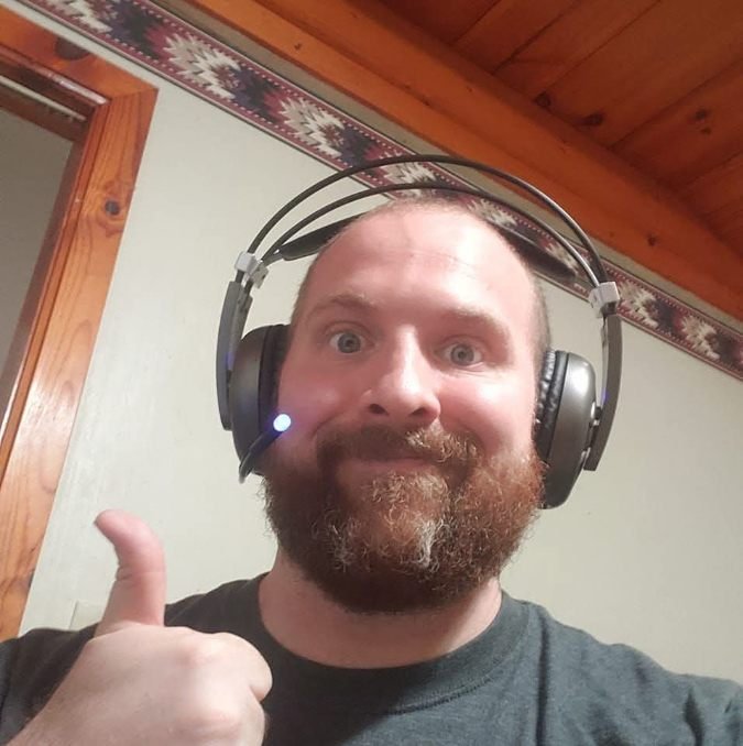 A man with a beard and short hair wearing a headset with a microphone, giving a thumbs up and smiling at the camera inside a wooden-paneled room.