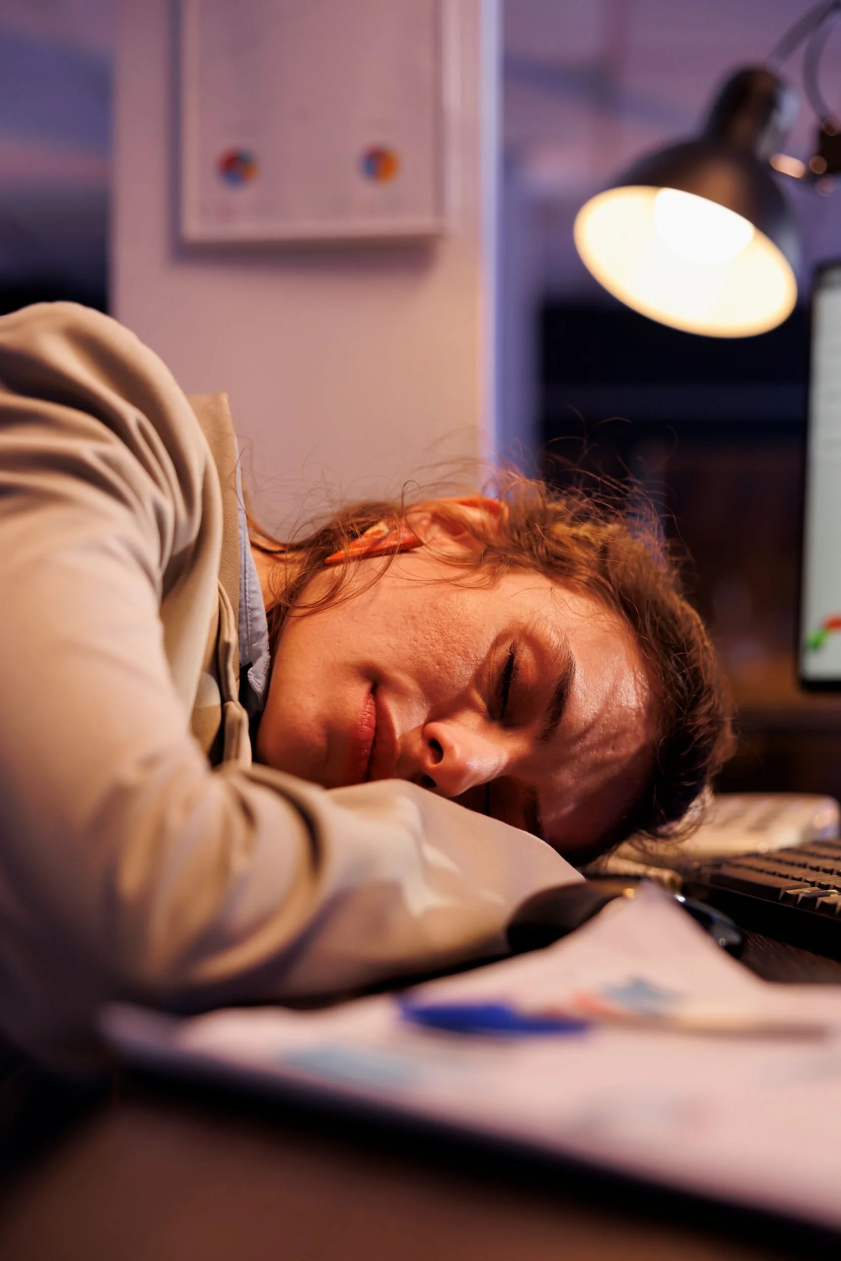 A young woman sleeping on her desk with her head resting on a computer keyboard, in a dimly lit room with a desk lamp and a computer monitor.
