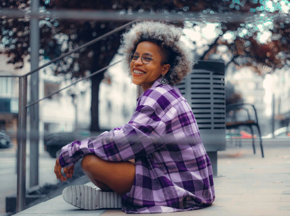 A young woman with curly gray hair, wearing glasses and a purple plaid shirt, sitting on the ground outdoors, smiling, with blurred trees and cityscape in the background.