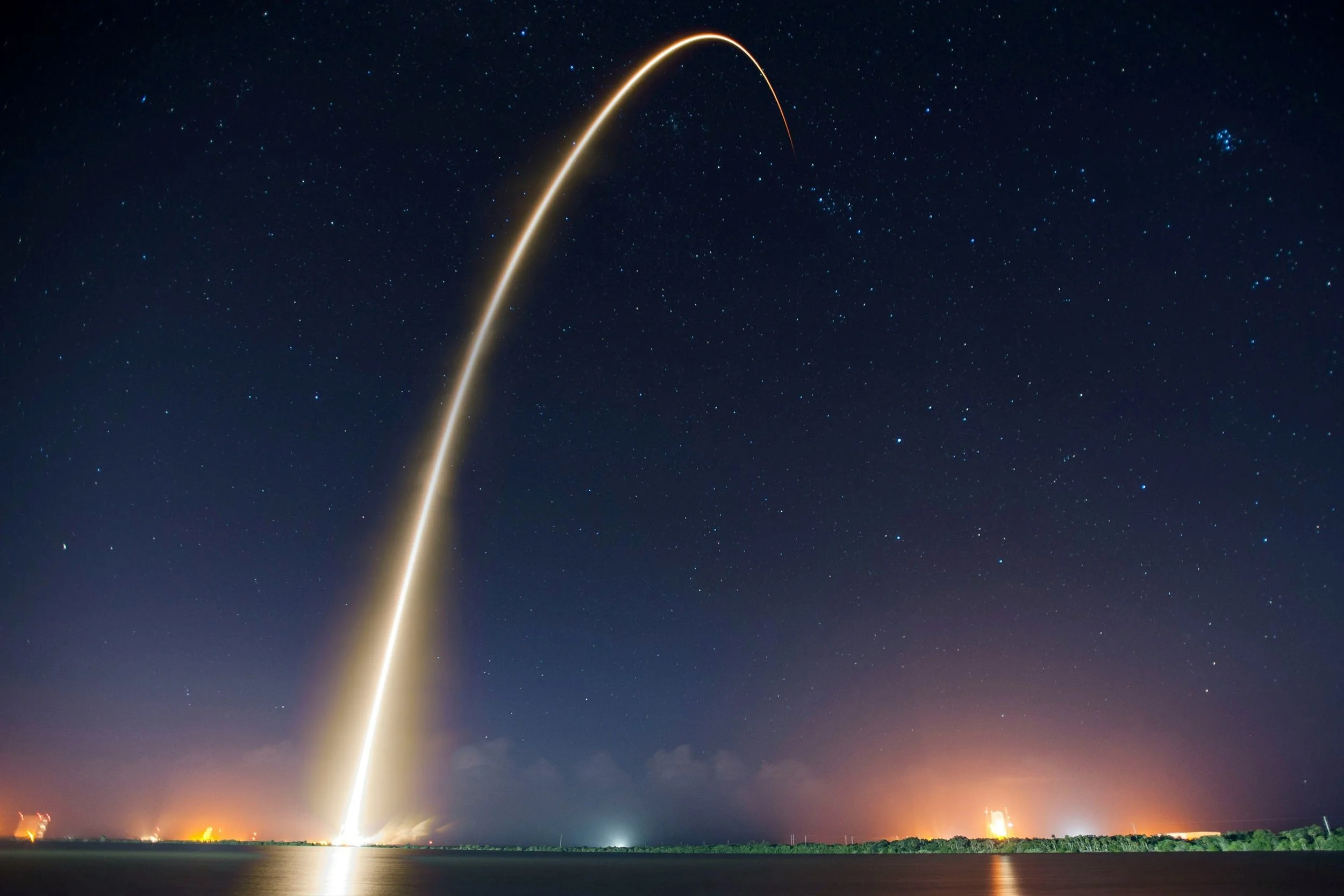 Rocket launch at night with bright trail streaking into the starry sky, reflecting on water below and city lights in the distance.