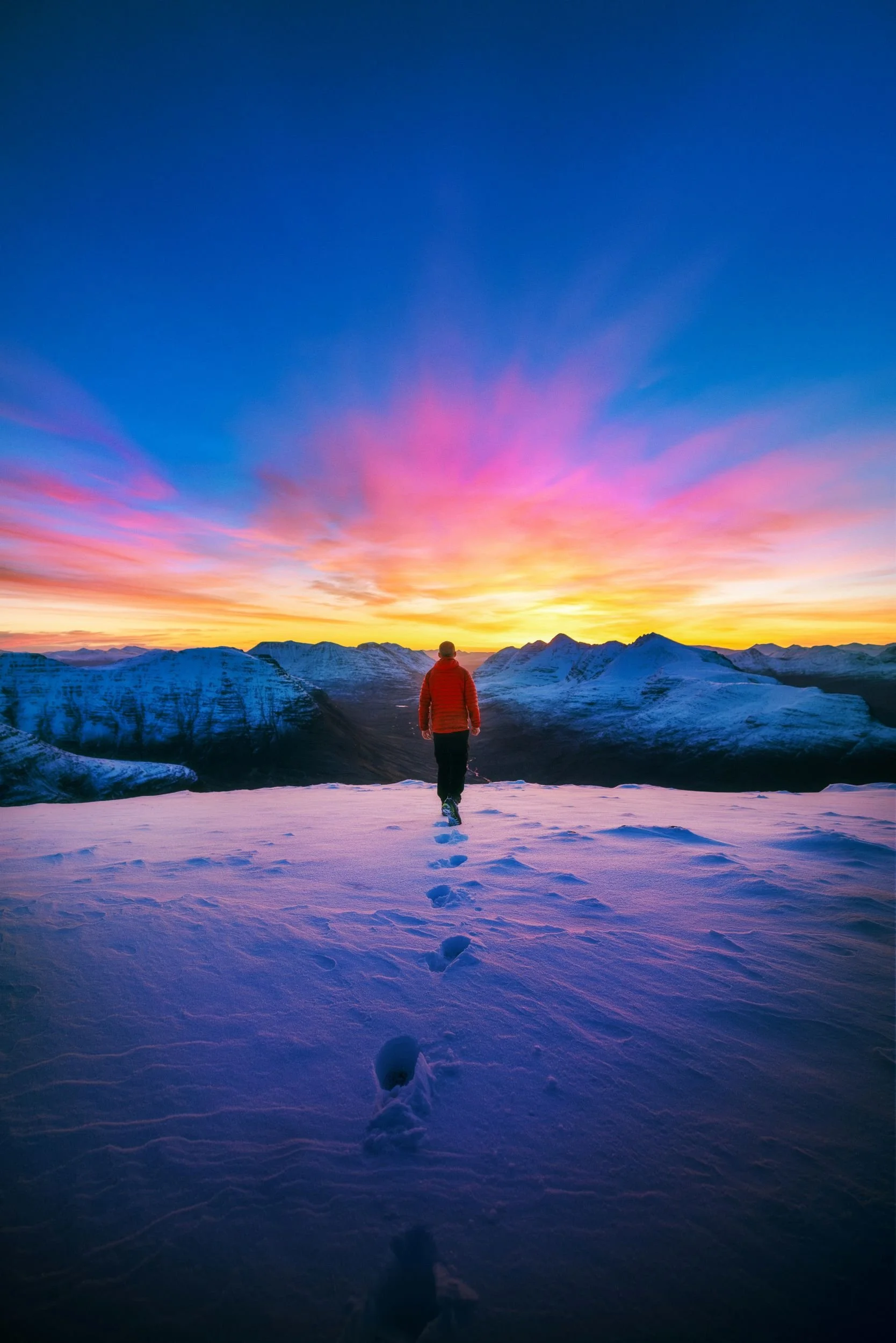 A person walking on snow-covered ground towards mountains at sunset, with a colorful sky of pink, purple, yellow, and blue hues.