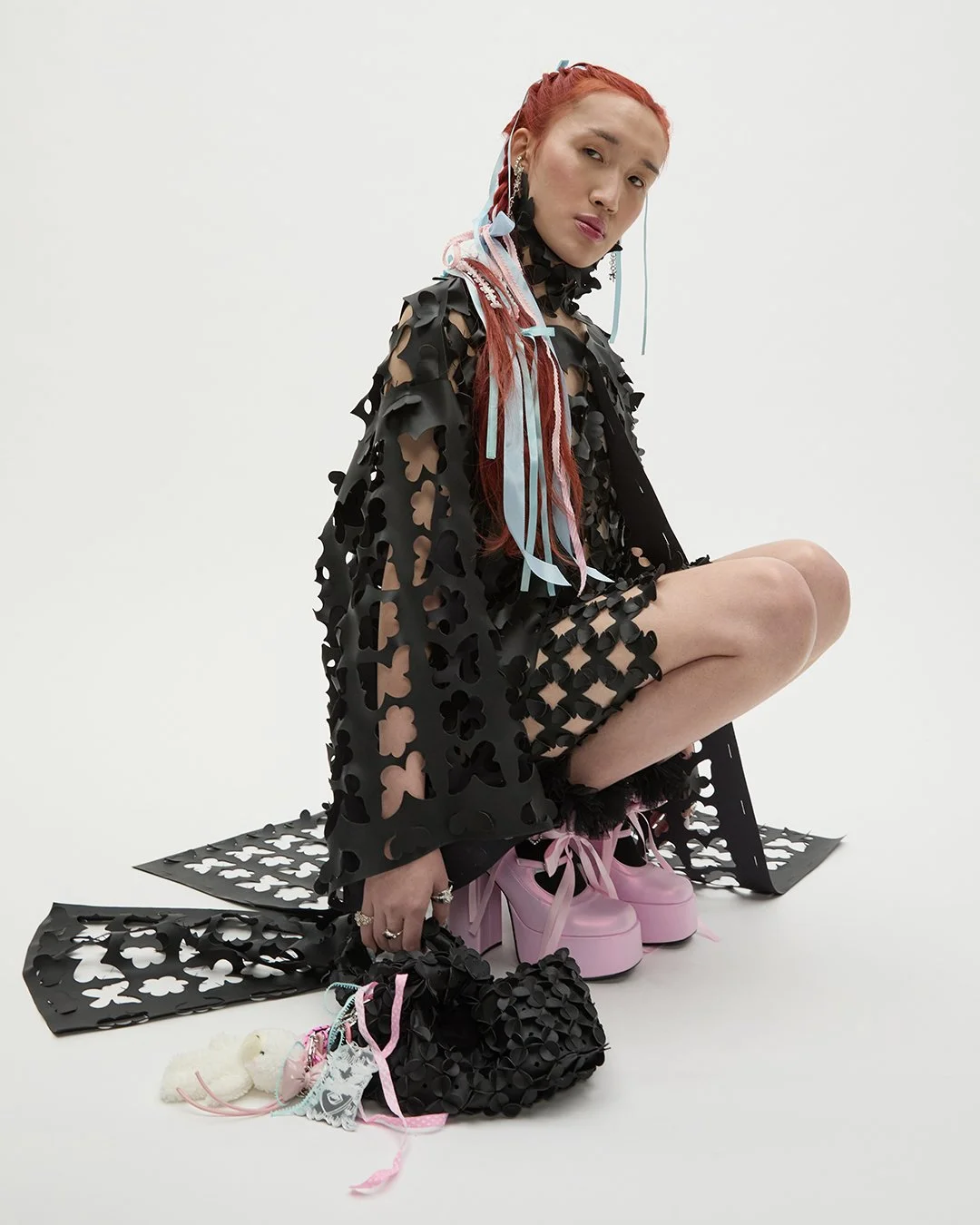 Junge Frau in schwarz mit ausgefallener Kleidung und pinken Schuhen, sitzt auf dem Boden vor weißem Hintergrund, hält einen schwarzen Taschen mit Blumenmuster und trägt mehrere Ringe sowie große Ohrringe.