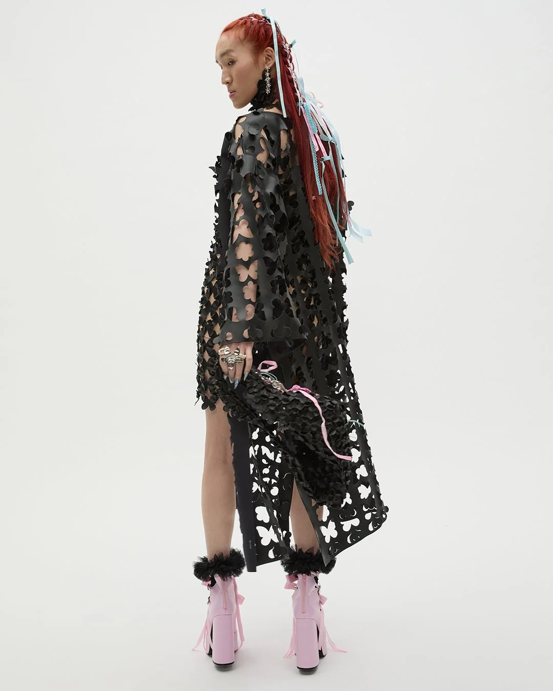 Frau trägt ein schwarzes Kleid mit Aufnähern in Herzform, pinke Schuhe mit Schleifen und trägt eine Handtasche aus ähnlichem Material, hält mehrere Ringe und hat rote geflochtene Haare mit bunten Bändern.