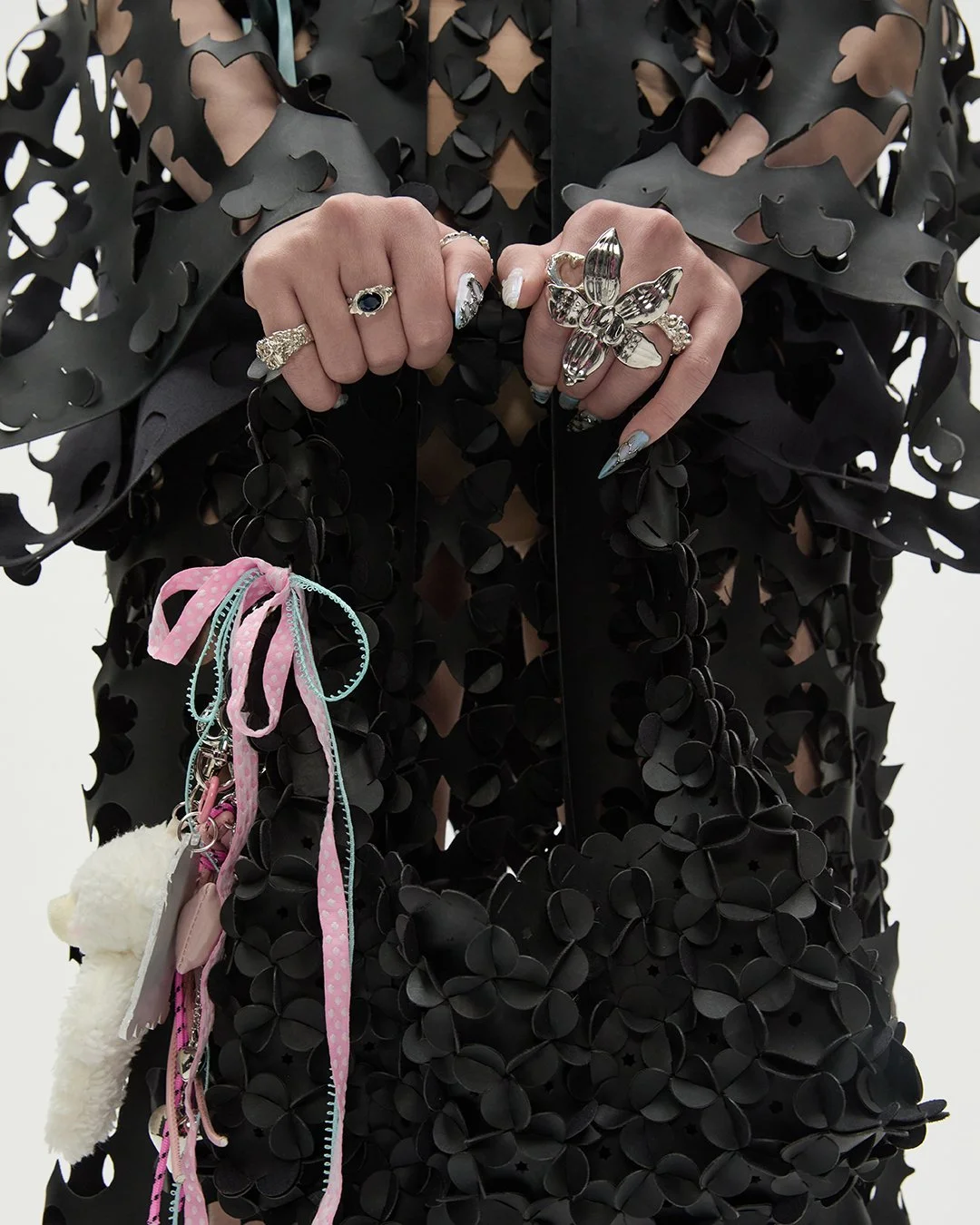 Nahaufnahme von Händen mit mehreren Ringen, in einem schwarzen Outfit mit Blütenmuster, die Hände halten eine schwarze Tasche mit seitlich angehängten, pinken, blauen und weißen Schleifen und einem kleinen weißen Plüsch-Anhänger.