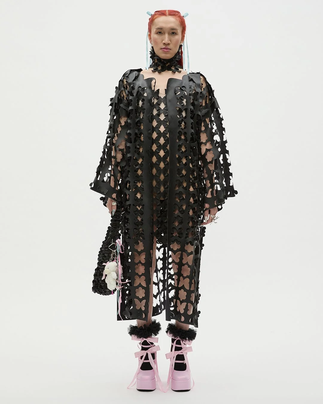 Frau in auffälligem schwarzen Kleid mit Lochmuster, pinken Plateauschuhen und buntem Accessoire-Behälter, vor weißem Hintergrund.