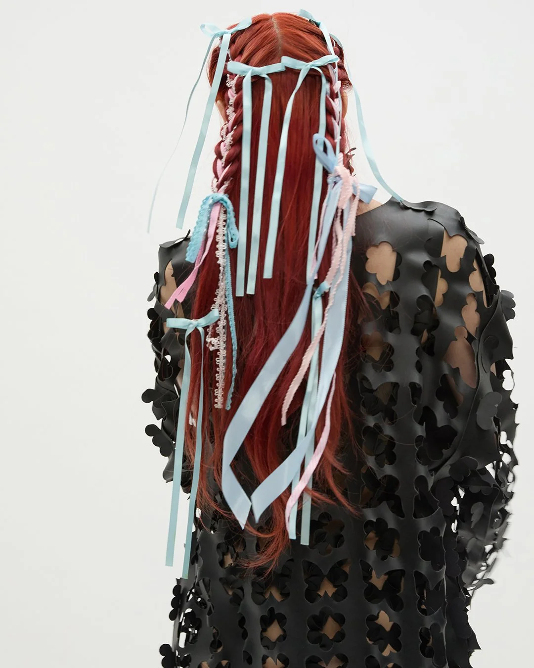 Eine Person mit roten Haaren, deren Rücken zur Kamera zeigt, trägt eine schwarze Jacke mit cut-out Blumenmuster und hat lange, verzierte Zöpfe mit blauen, rosa und weißen Bändern und Haargummis. Der Hintergrund ist schlicht und hell.