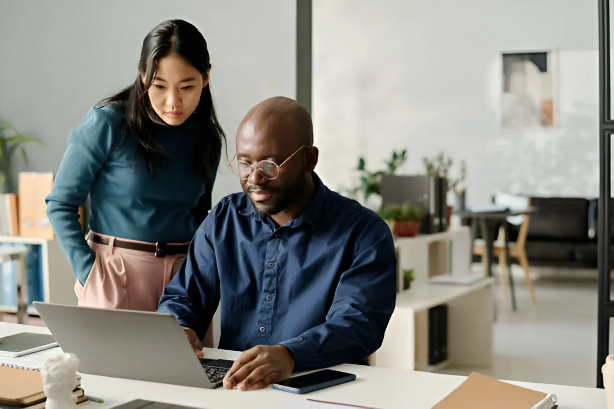 Two colleagues collaborating at a desk with a laptop in an office setting.