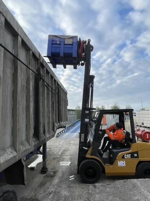 A forklift operator in an orange safety vest and black hat lifting a blue crate on a black forklift near a large gray container on a dock or industrial site.
