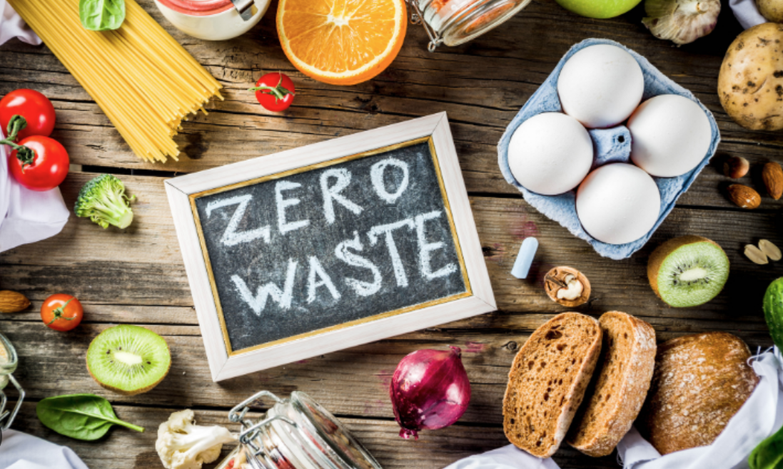 Assorted fresh fruits, vegetables, eggs, bread, and pasta on a wooden table with a chalkboard that reads 'ZERO WASTE'.